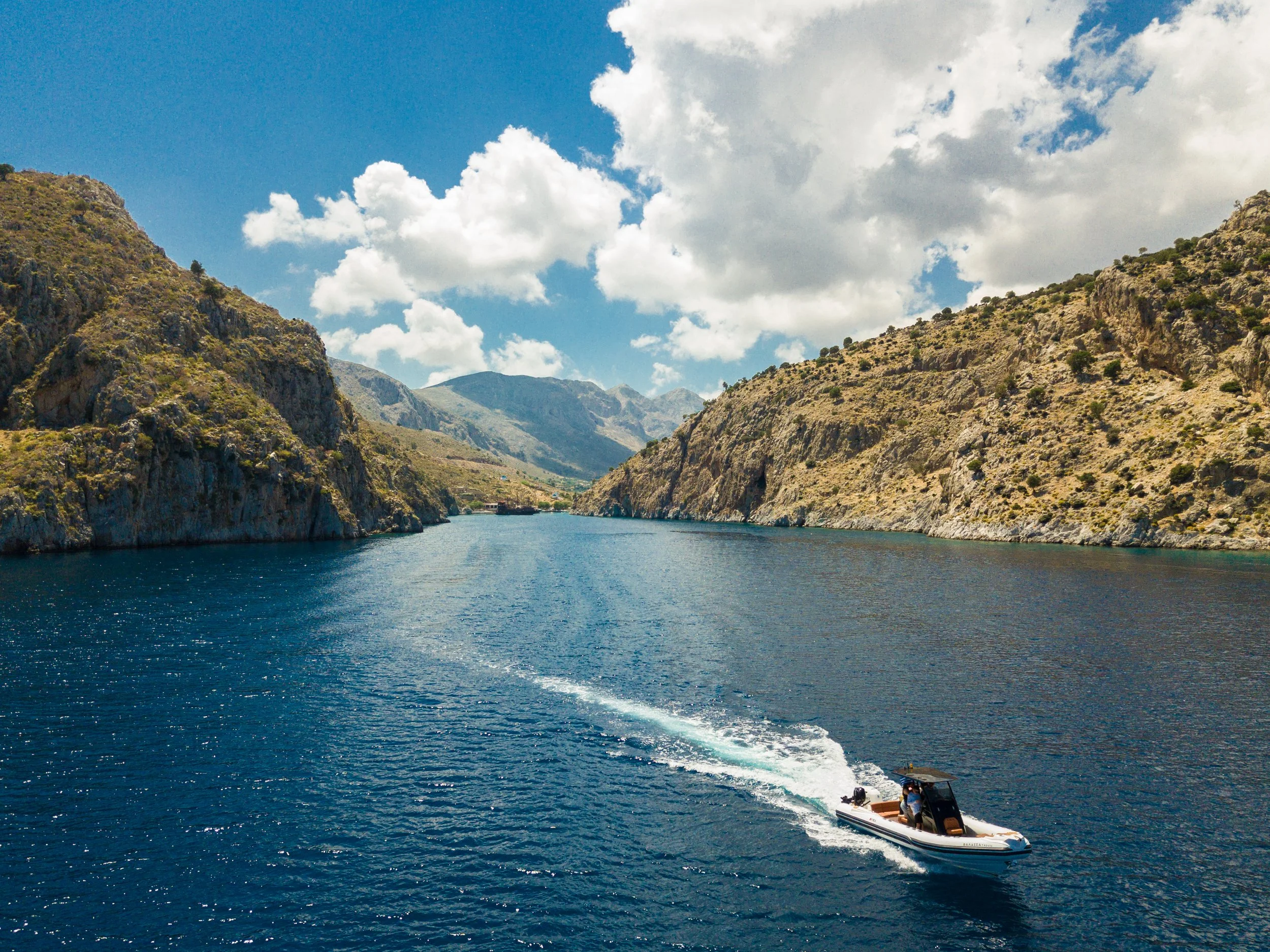 A boat riding through a deep blue river between rugged, desert-like mountains with sparse vegetation. The sky is partly cloudy with large, fluffy clouds.