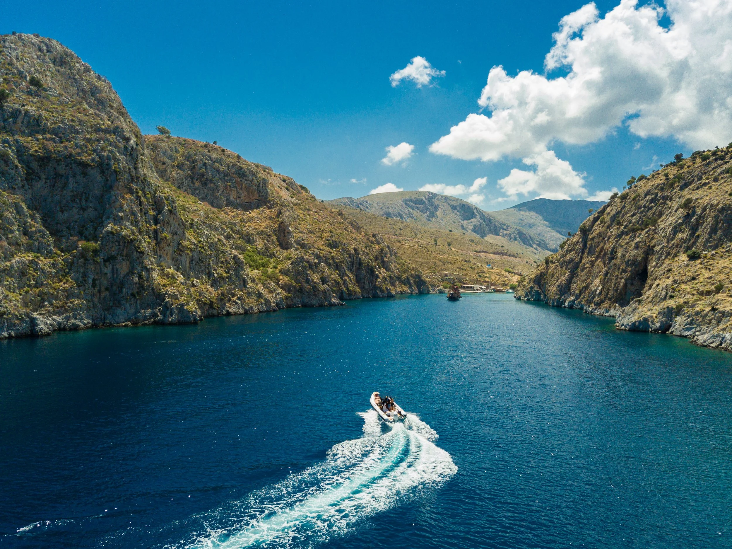 A boat speeding through a narrow canyon with steep rocky cliffs on either side and a clear blue sky with some clouds above.