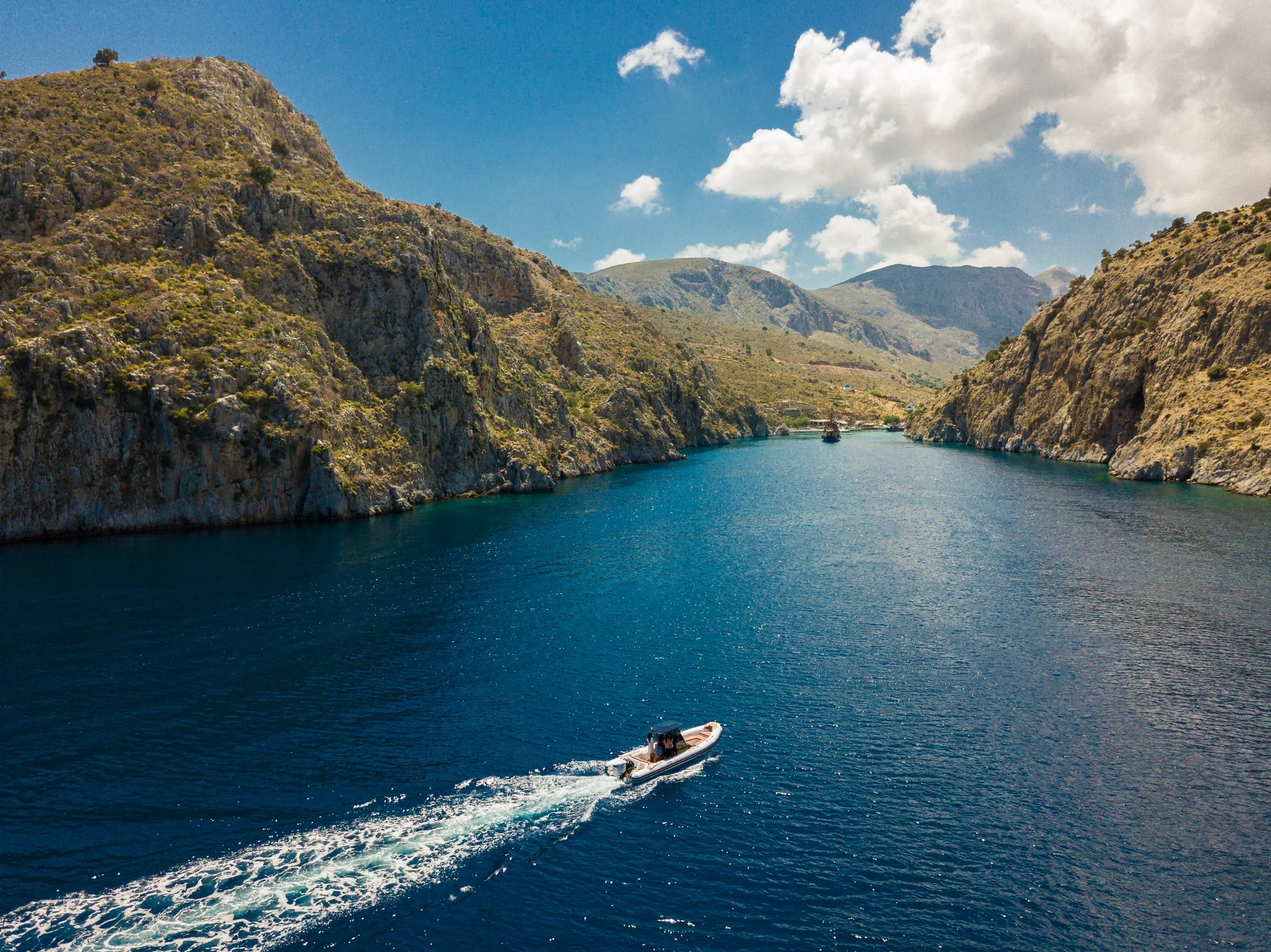 A small motorboat with people on board traveling through clear blue water in a river surrounded by brown rocky mountains under a partly cloudy sky.