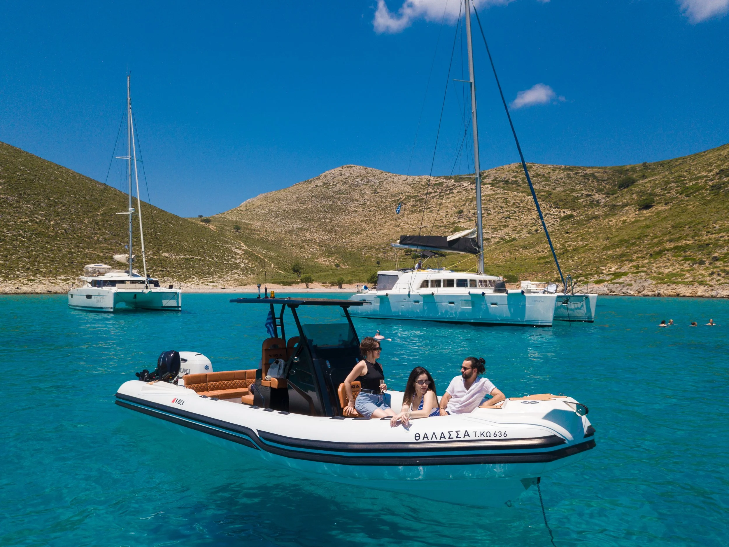 People sitting in a boat with a sailboat and mountainous island in the background, under a blue sky with some clouds.