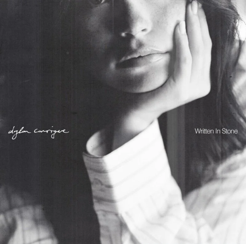 Black and white close-up photo of a woman's face, resting her head on her hand, with handwritten text reading "dylan cornoge" and printed text "Written In Stone."