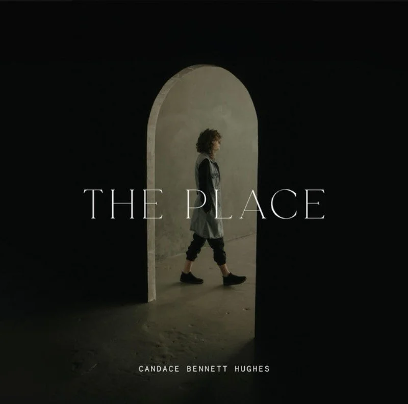 A woman with curly hair walking through an arched doorway, with a dark background and a light-colored wall inside. The text 'The Place' and 'Candace Bennett Hughes' are overlaid on the scene.