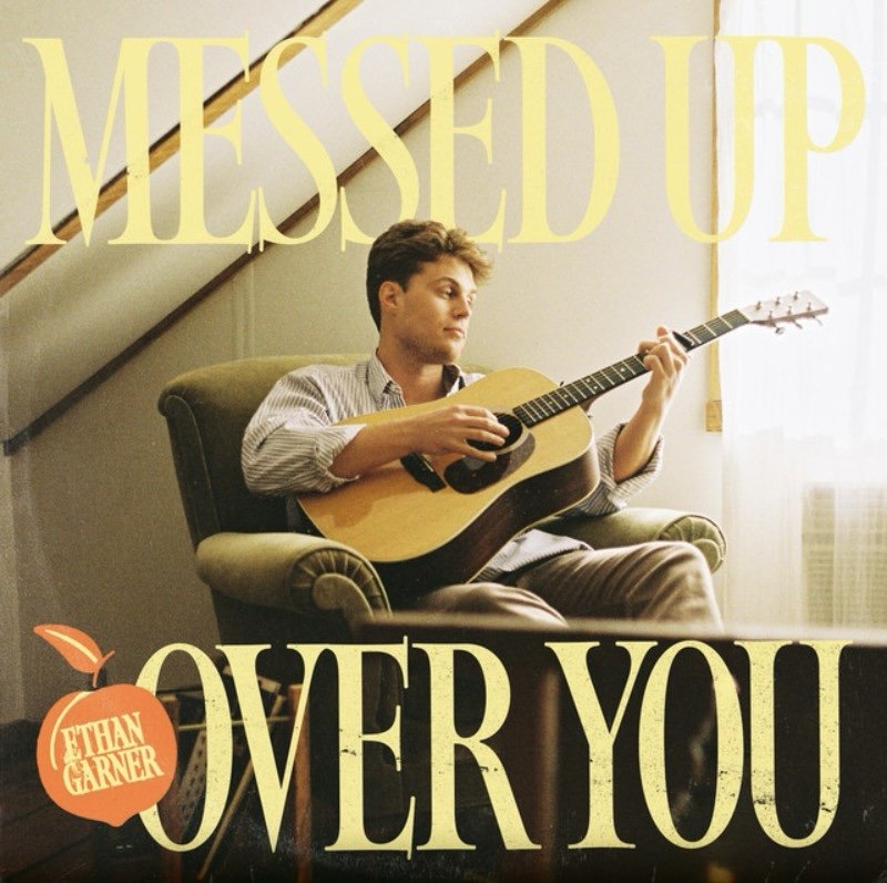 Young man sitting in a chair playing an acoustic guitar, with large yellow text reading 'Messaged Up Over You' and an orange apple graphic with the text 'Ethan Garner' on it.