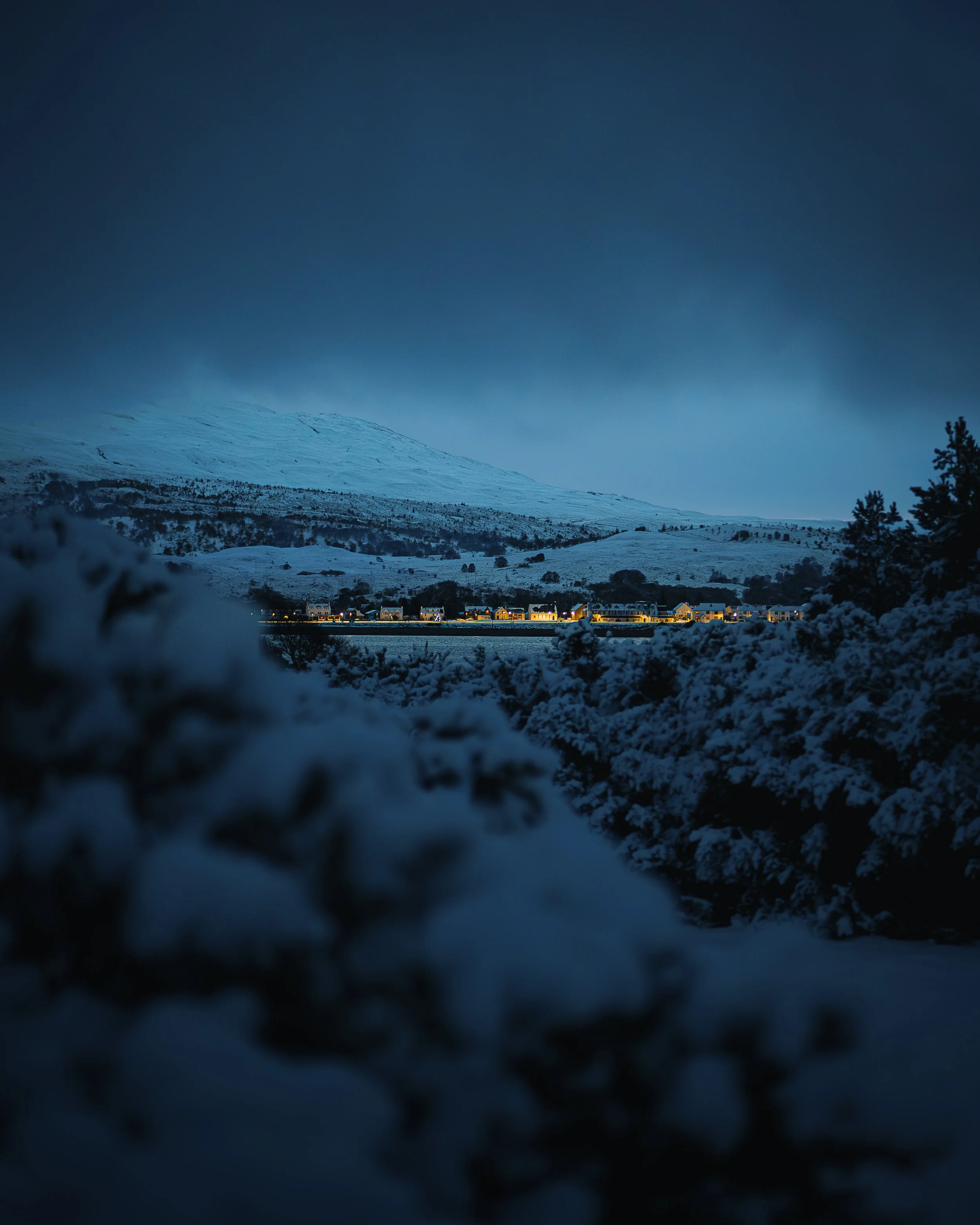 Snow-covered landscape with a row of houses illuminated at night, distant mountain under dark cloudy sky.