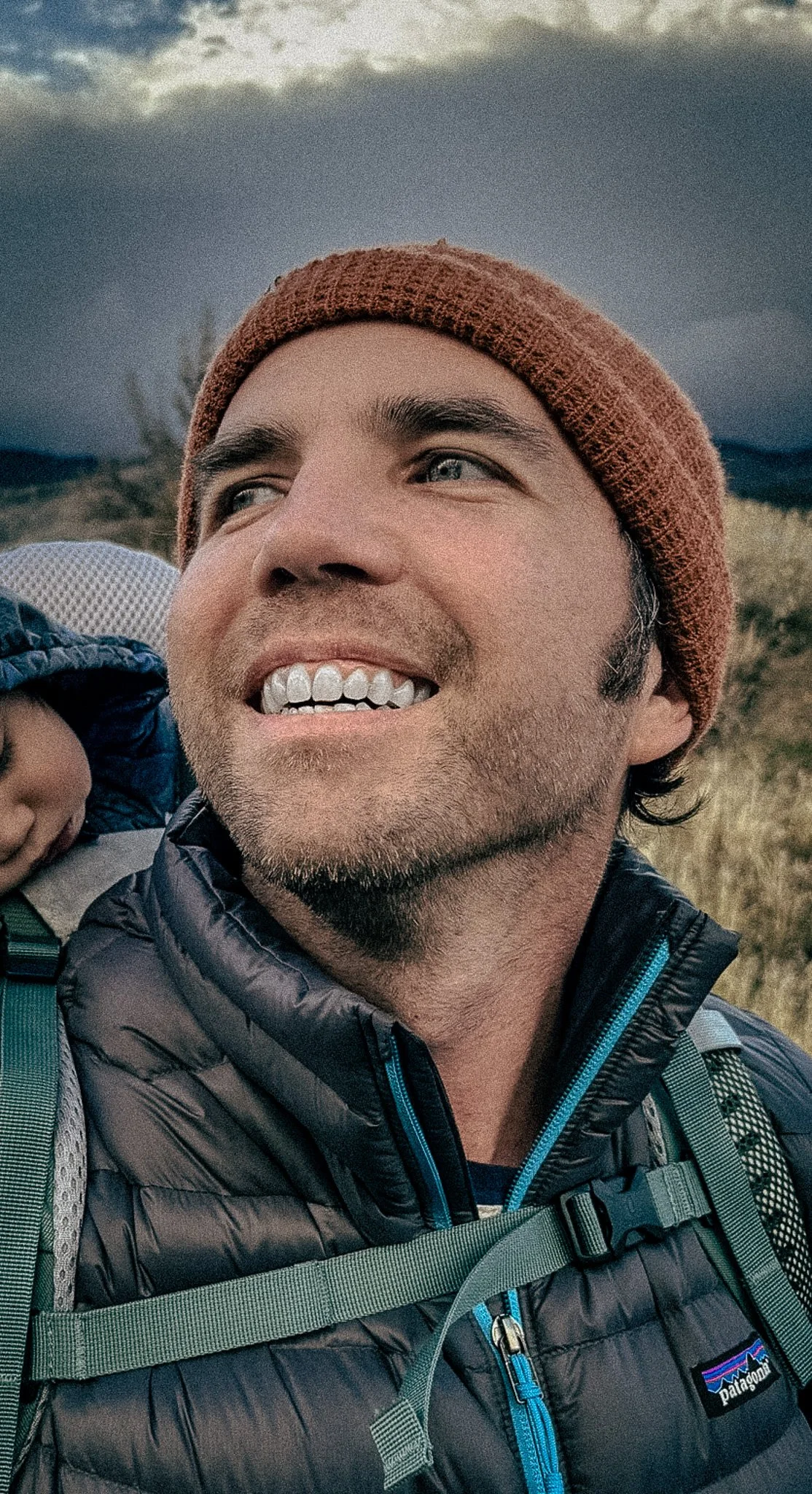 Close-up of a smiling man wearing a brown beanie and a black Patagonia puffer jacket, with a child on his back, outdoors with cloudy sky and open landscape in the background.