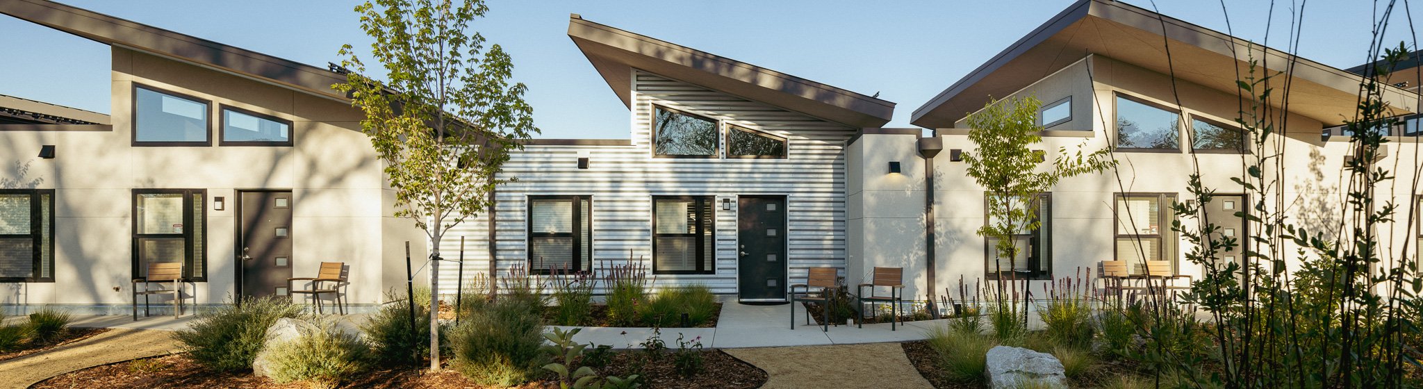 Modern row of residential townhouses with angled roofs, large windows, trees, benches, and landscaped front yards.