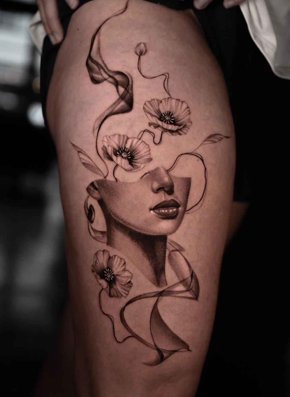 Black and gray tattoo of a woman's face with flowers and abstract lines, placed on the upper thigh.