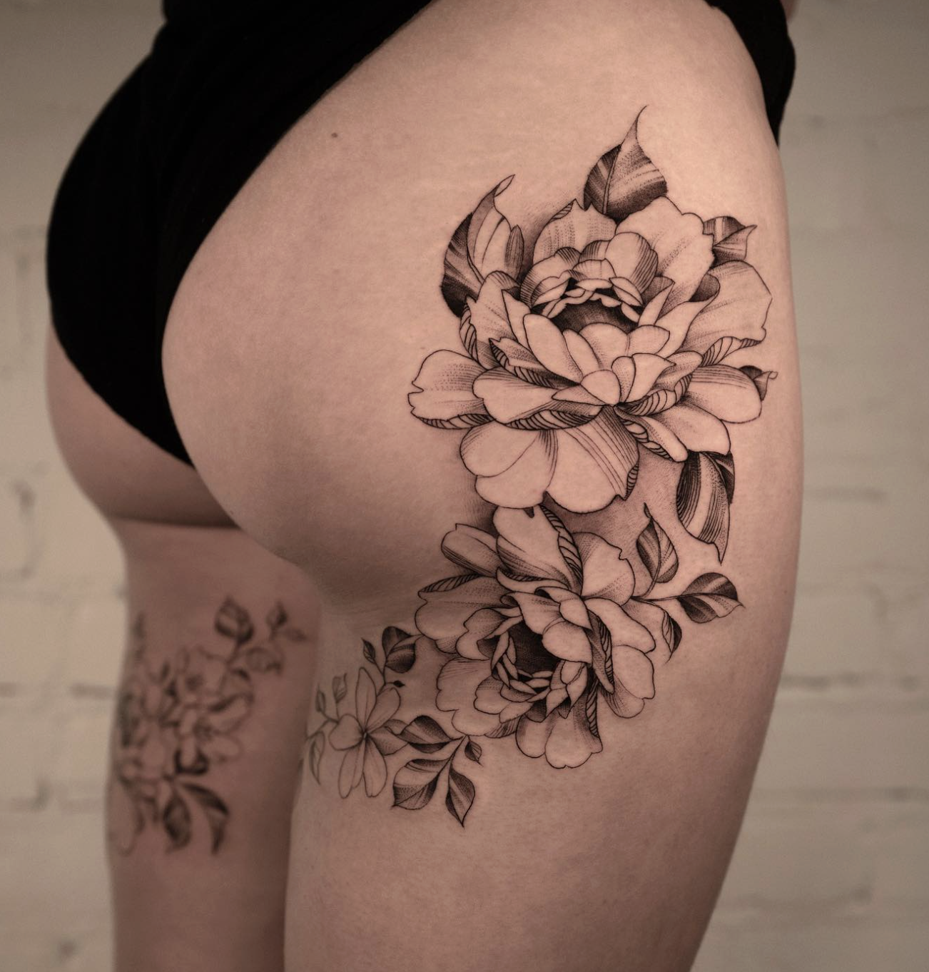 Close-up of a black and gray floral tattoo on a person's shoulder and upper arm, featuring a large blooming flower with detailed petals and leaves.