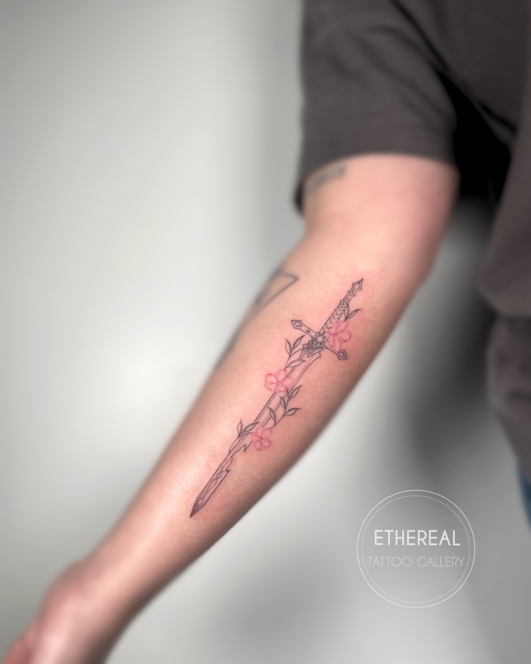 Tattoo of a sword with pink flowers and leaves on a person's forearm.