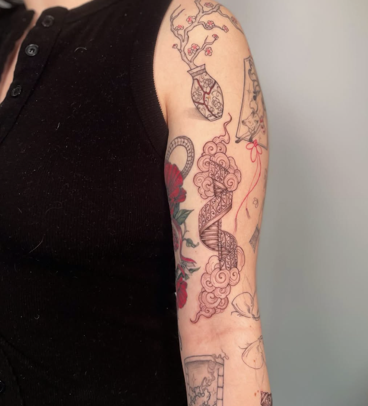 A person's upper arm with several tattoos, including a spiral stairway, stylized clouds, a cherry blossom tree in a vase, a snake, and various smaller designs, all in black ink with some red accents.