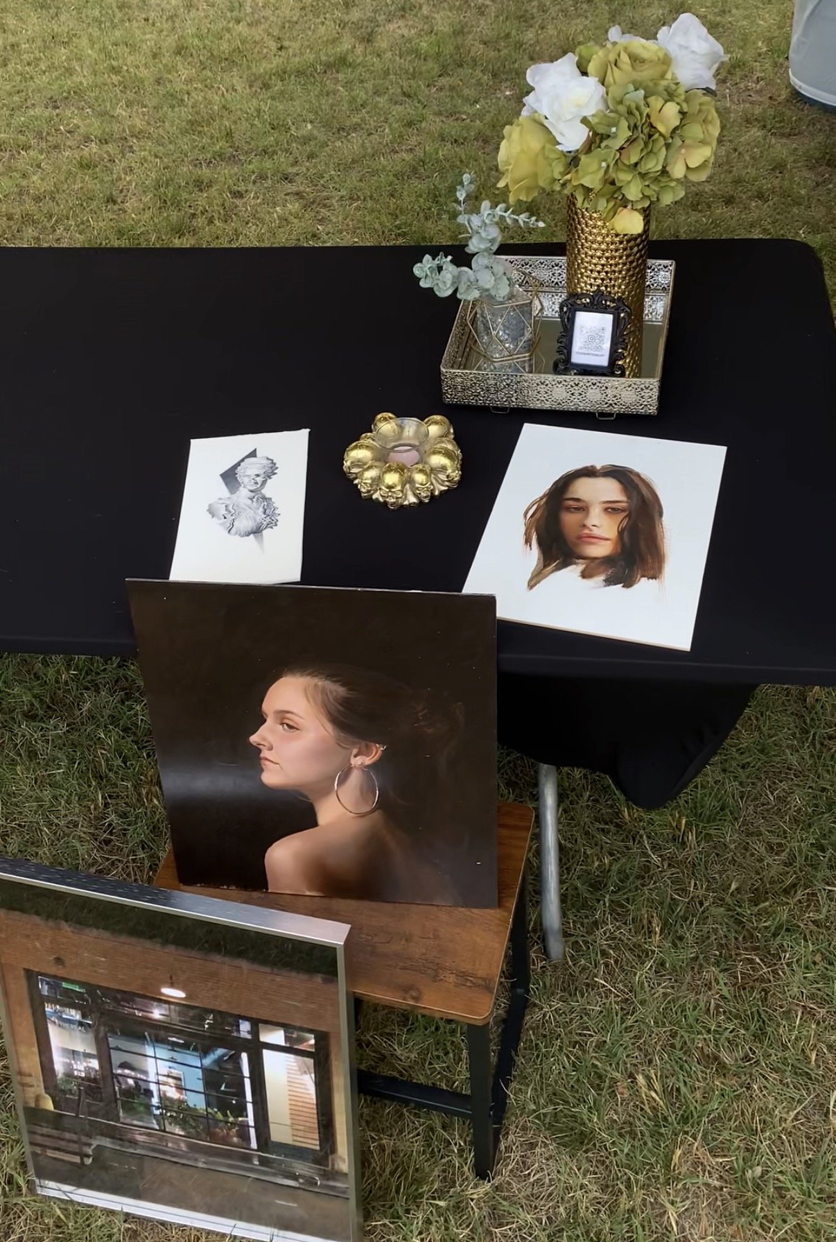 A photo display booth with framed photographs of women, a bouquet of yellow and white flowers, and decorative items on a black table, set outdoors on grass.