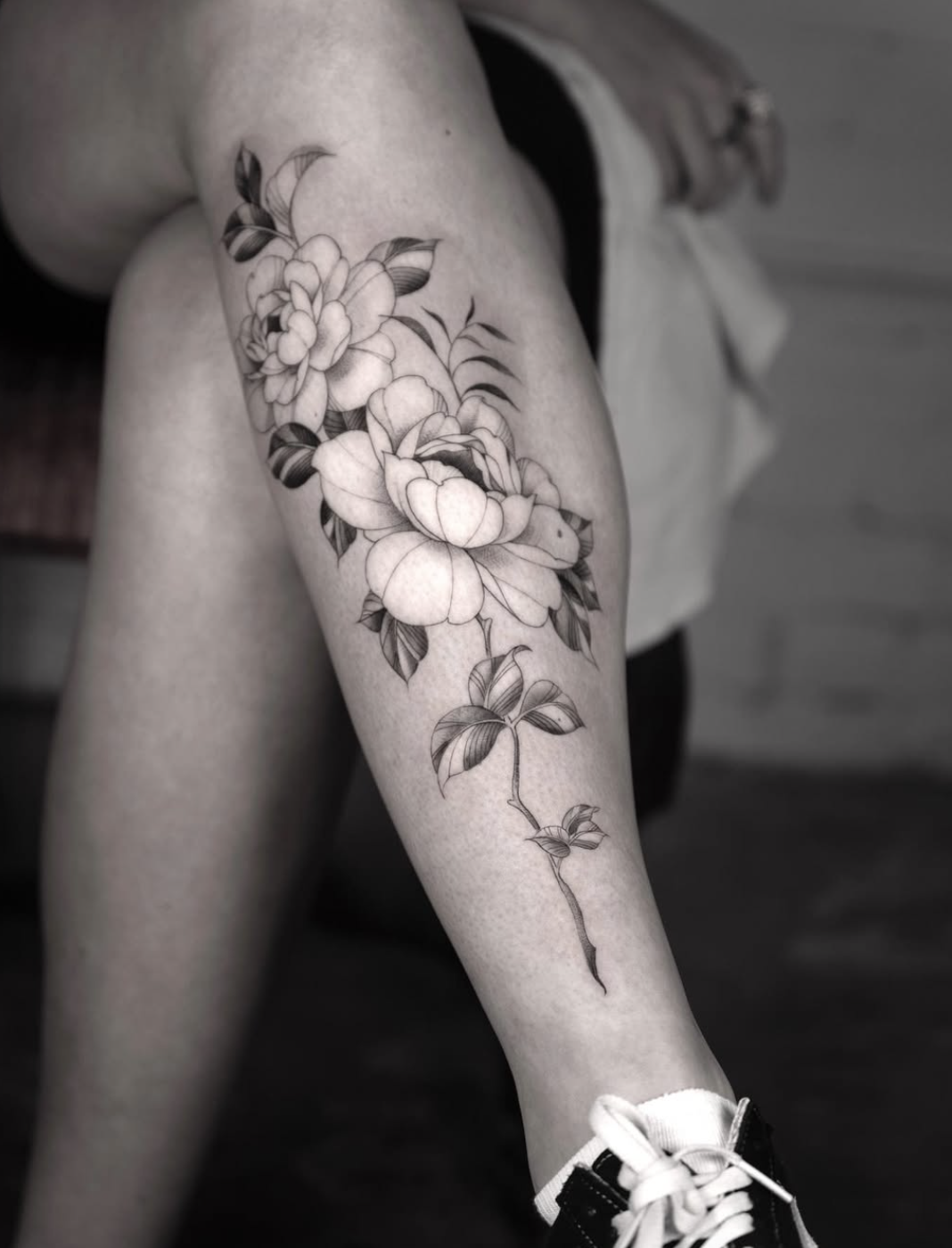 Black and white photo of a person's thigh with a detailed floral tattoo of a large flower, leaves, and a stem.