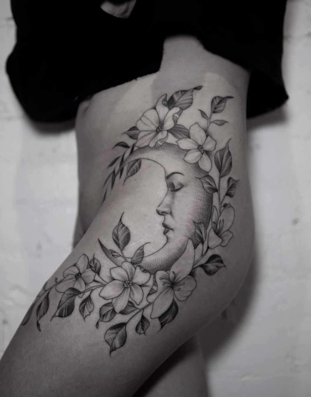 Black and gray tattoo of a crescent moon with a woman's face in profile, surrounded by detailed flowers and leaves, on a person's thigh.