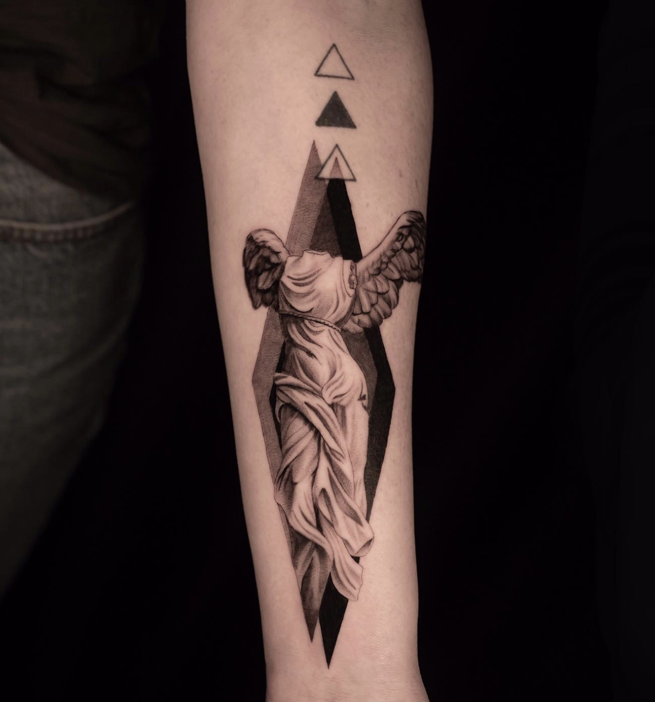 Tattoo of a classical statue with wings, draped fabric, and geometric shapes above it, on a person's forearm.