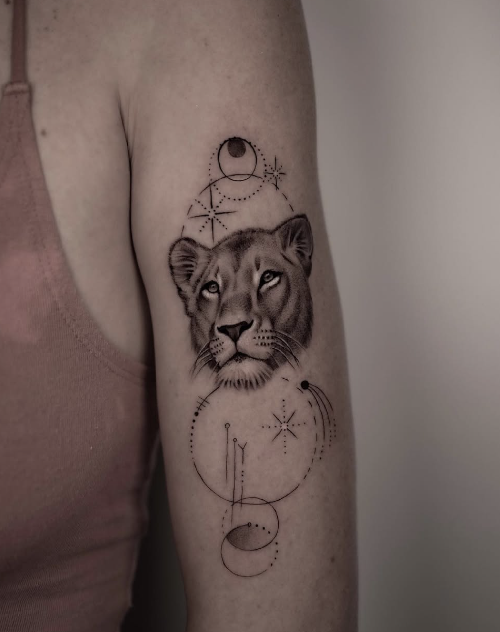 Tattoo of a lioness's face surrounded by mystical celestial and geometric designs on a person's upper arm.