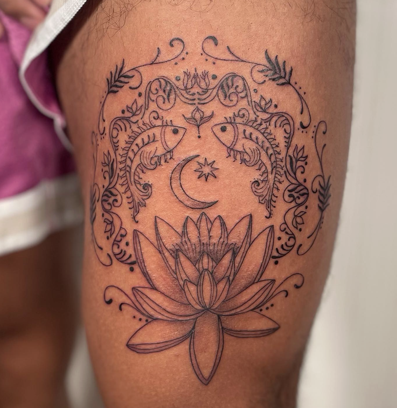 A detailed tattoo on a person's arm featuring a lotus flower at the bottom, a crescent moon, a star, two fish, and decorative floral and scroll patterns.