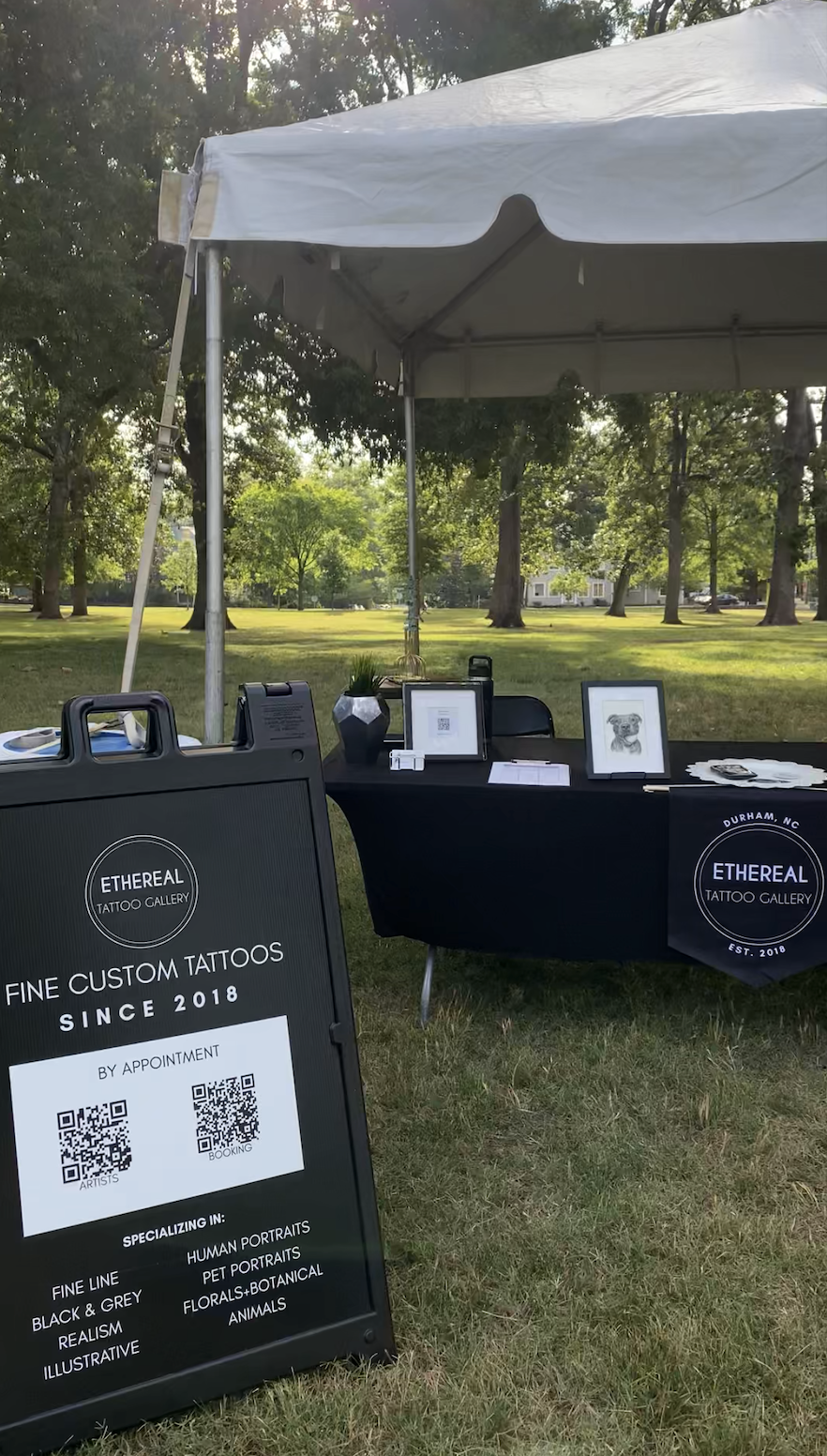 Outdoor art booth under a white canopy tent, displaying framed artwork, a decorative plant, and promotional materials for Ethereal Tattoo Gallery in Durham, NC, set in a park with green grass and trees.