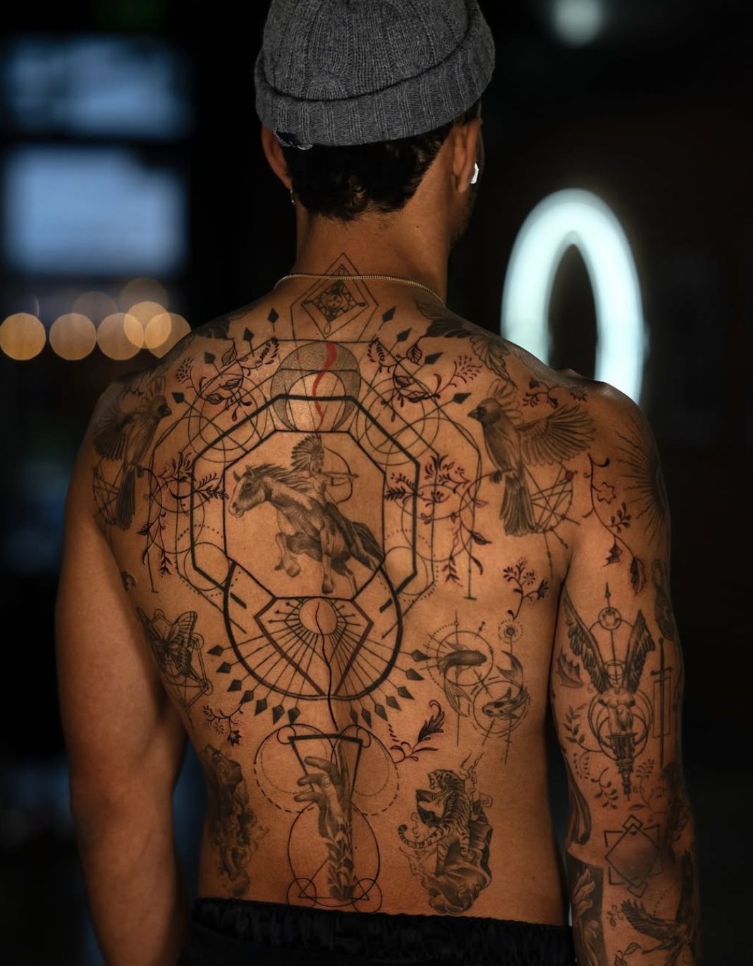 Back view of a person with numerous black tattoos on the back, shoulders, and arms, including geometric designs, animals, and symbols, wearing a gray beanie and white earbuds.