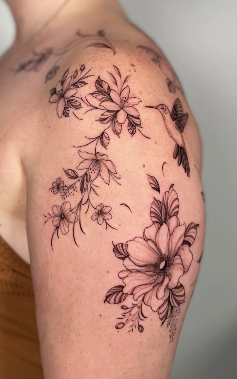 Black outline floral tattoo with large flowers, leaves, and a hummingbird on a person's shoulder and upper arm.