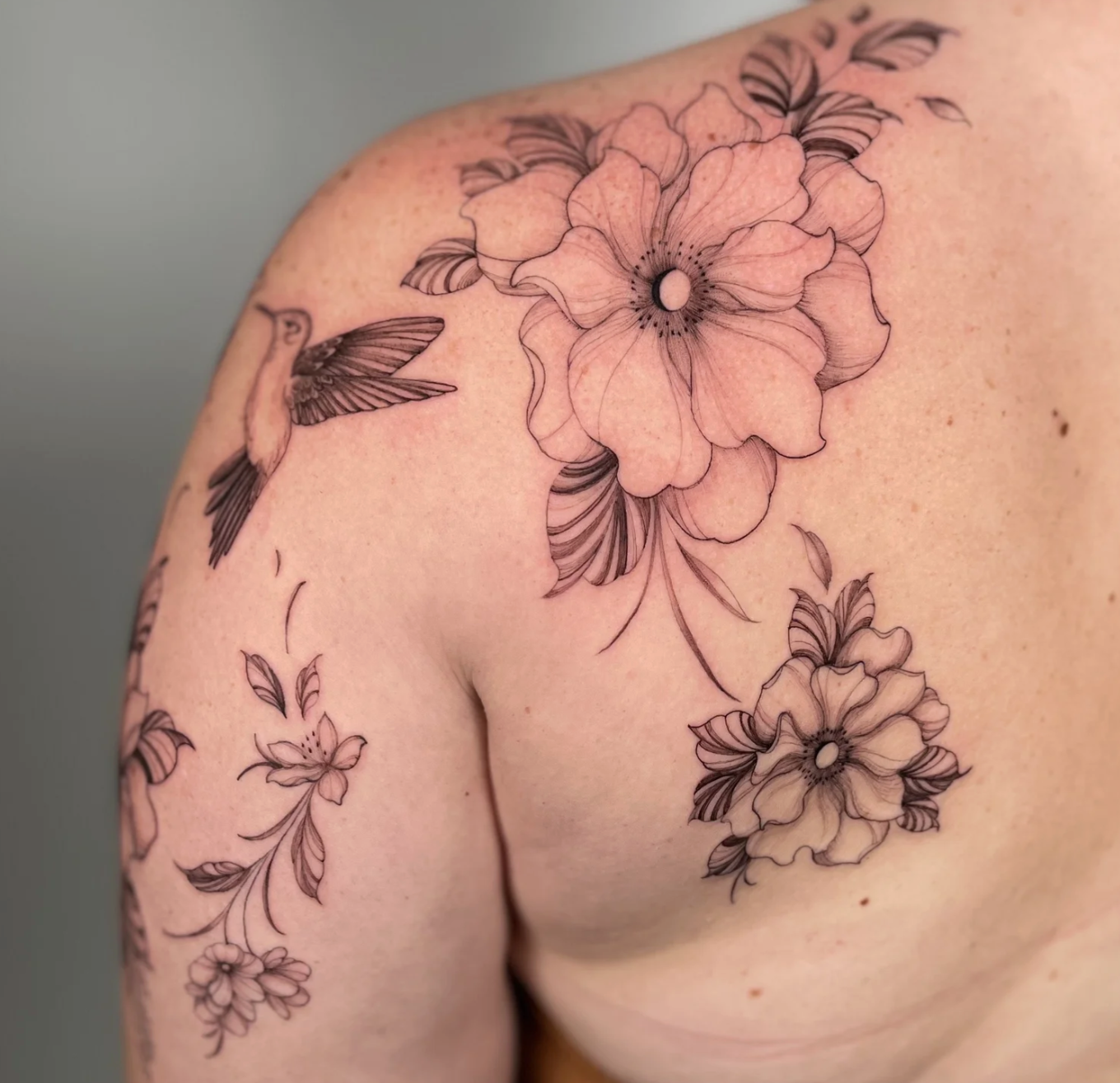 Black and gray floral tattoo with large flowers, leaves, and a hummingbird on a person's shoulder and upper arm.