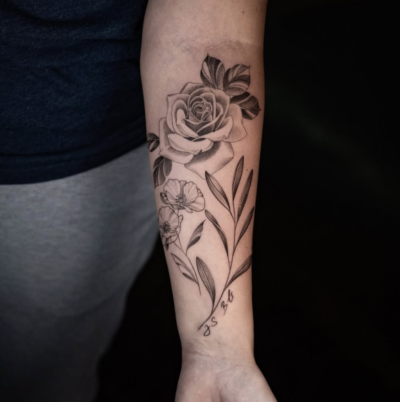 Black and gray tattoo of a rose with leaves, smaller flowers, and grass on a person's forearm, with initials 'J S B' written at the bottom.