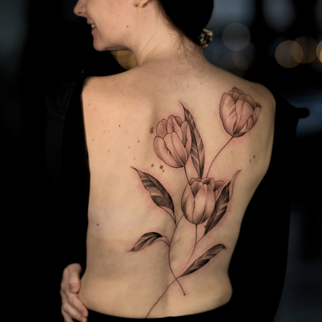 A woman with dark hair and a side profile view showing her shoulder and back, with a tattoo of pink flowers and leaves on her upper back.