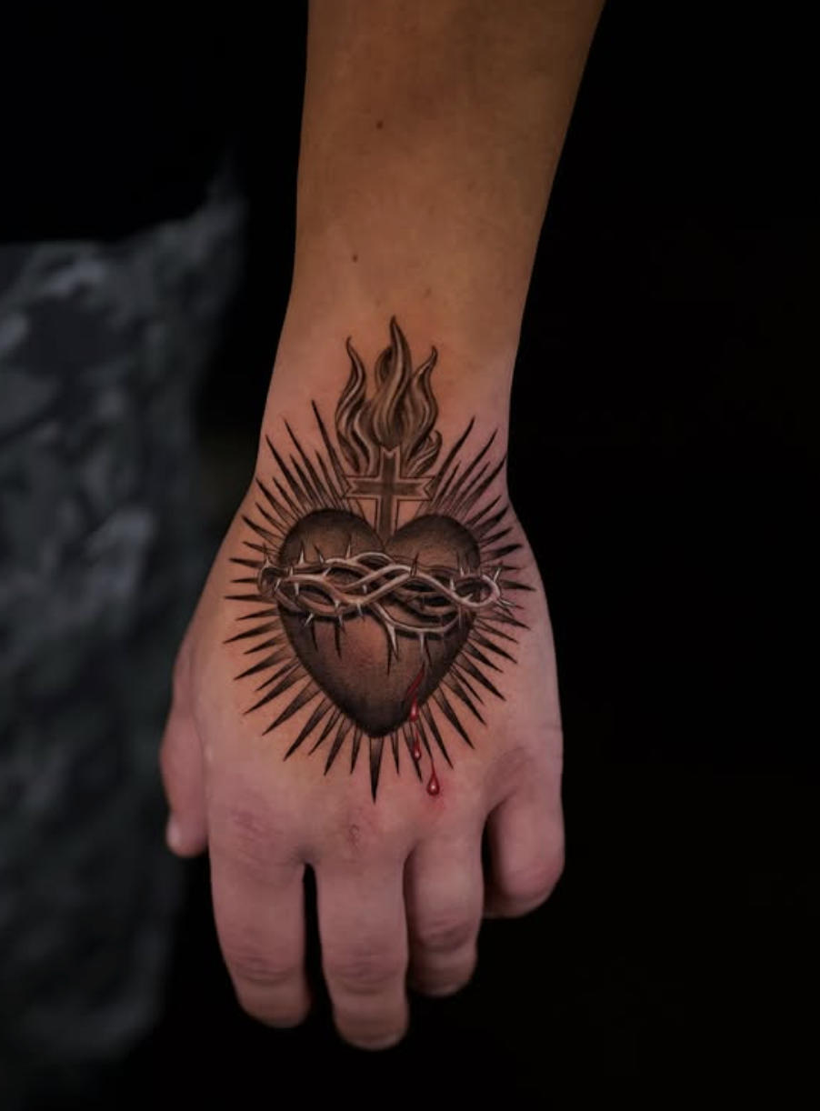 Fine line black and grey Sacred Heart hand tattoo with a flaming heart, crown of thorns, and cross, tattooed in realistic microrealism style on the back of the hand by Brian Parrillo at Ethereal Tattoo Gallery in Durham, North Carolina.
