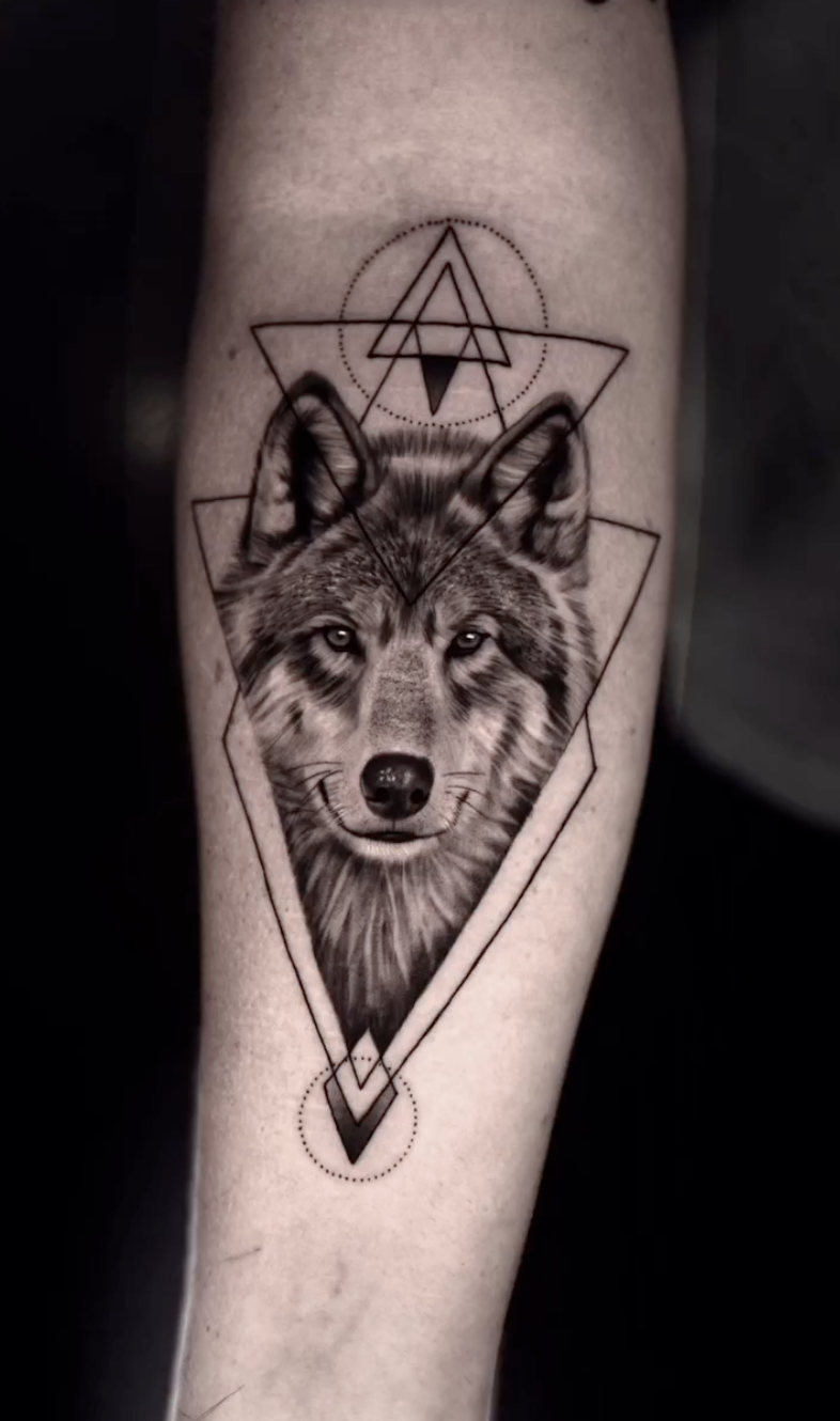 Tattoo of a wolf's face framed by geometric shapes and lines, with circles and triangles above and below.