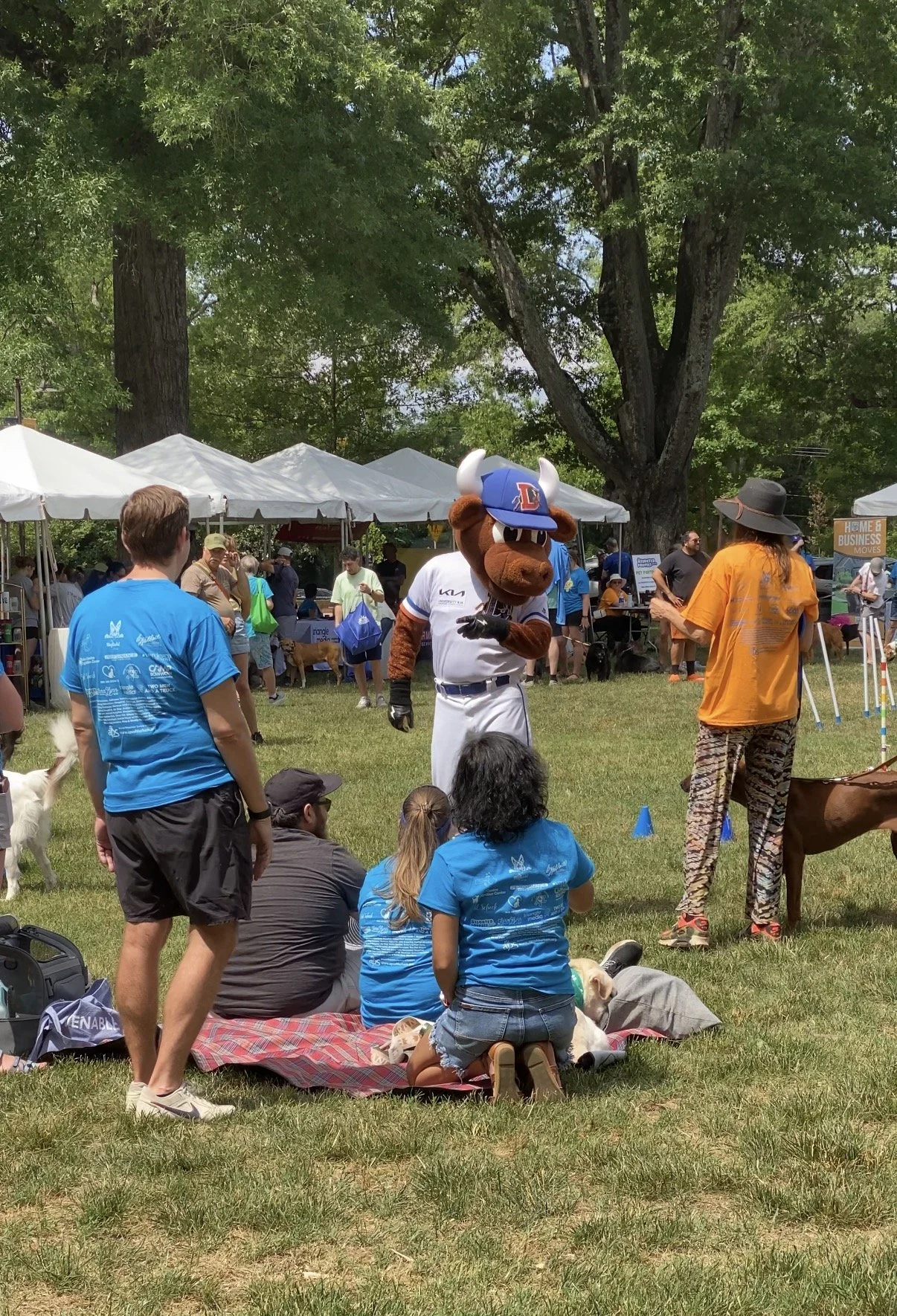 At an outdoor event, a person dressed in a sports-themed mascot costume of a dog in a baseball uniform and hat interacts with the audience, composed of children and adults, some sitting on a mat and others standing, under a large tree with white tents and booths in the background.