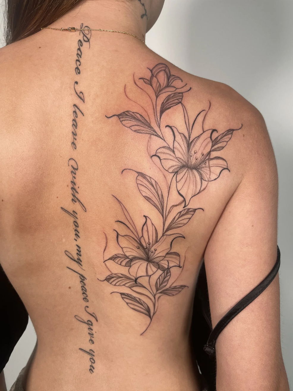 A person's back and shoulder with a large floral tattoo of lilies and leaves, and cursive text running vertically along the shoulder blade.