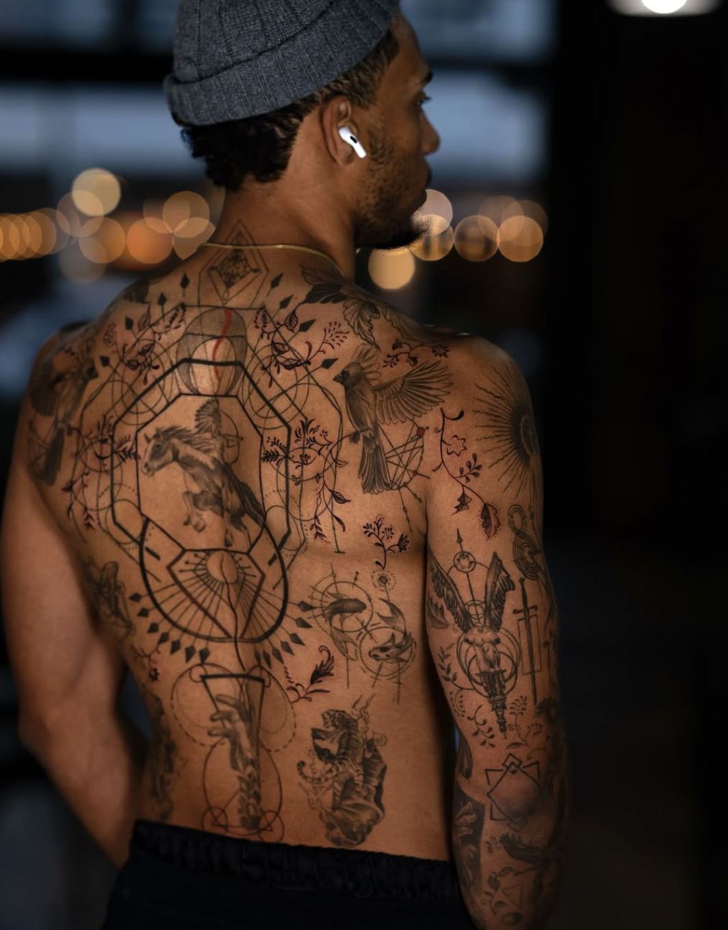 A shirtless man with tattoos on his chest and arm, wearing a gray beanie and white earphones, standing outdoors at night with blurred city lights in the background.