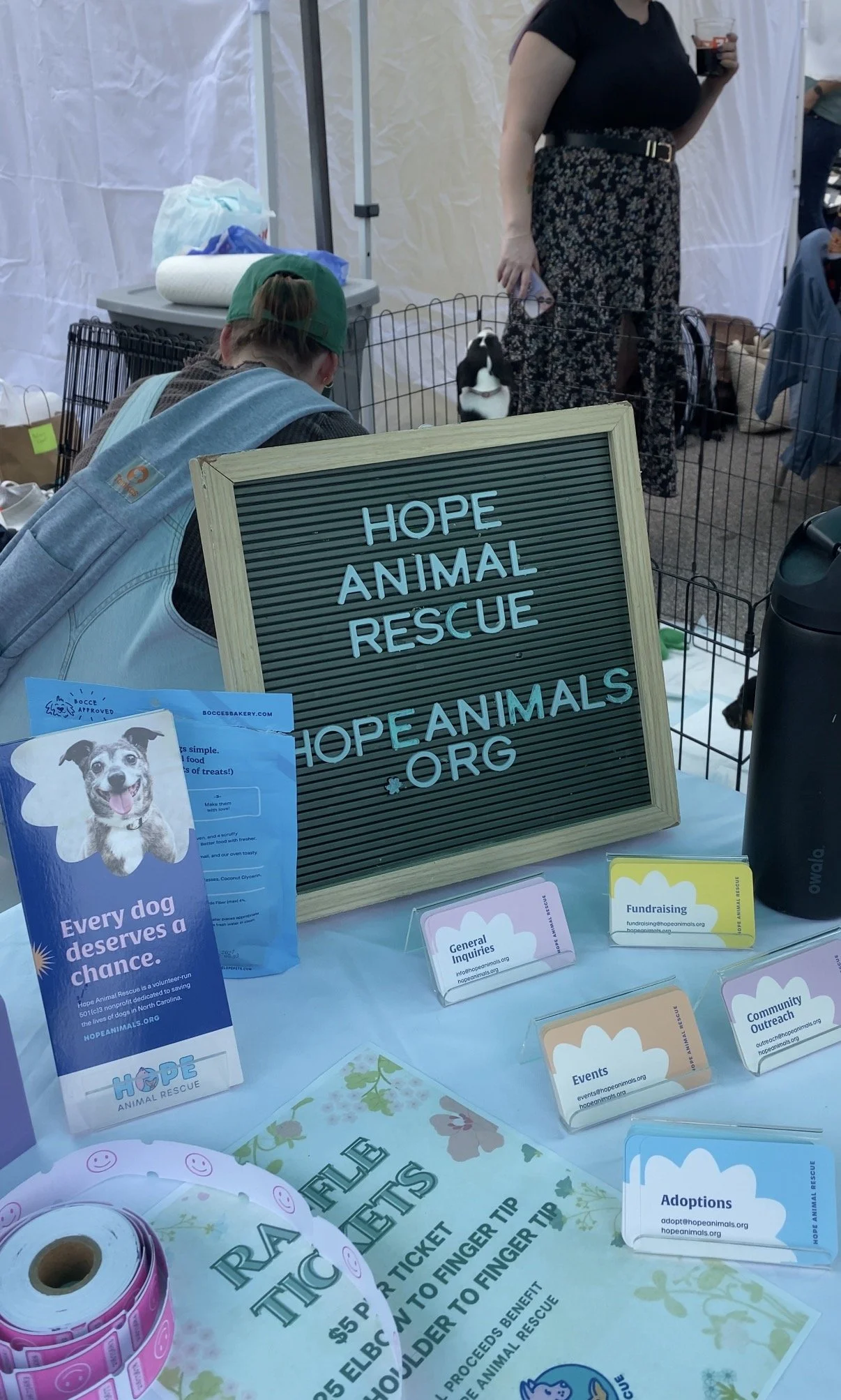 A table display at a Hope Animal Rescue booth features informational brochures, a framed sign with the organization name and website, and pet adoption materials. Behind the table, a woman in black and patterned pants stands near a small animal enclosure, with a person in a green cap and backpack sitting nearby.