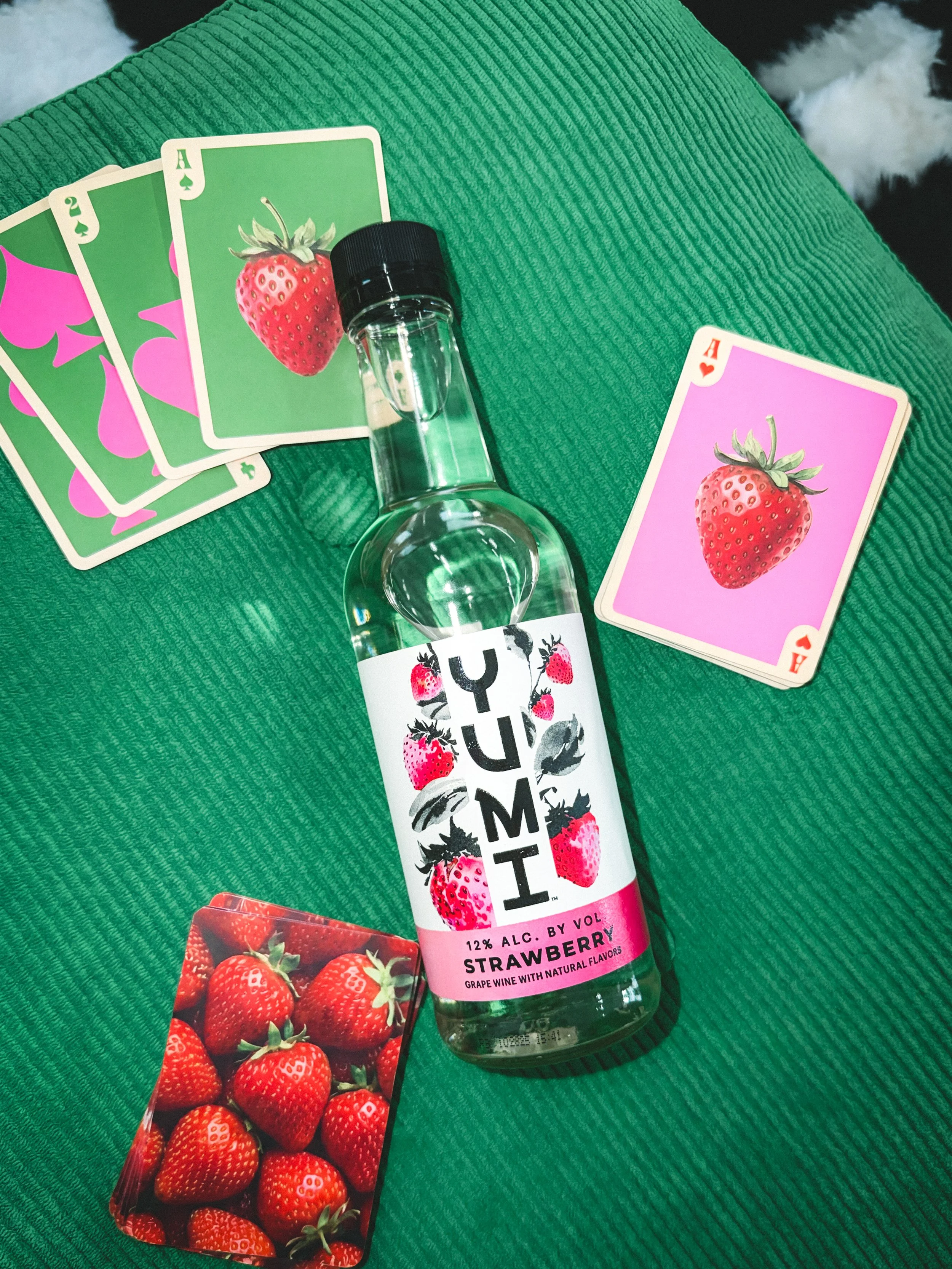 A glass bottle of YUMI strawberry flavored wine with strawberry illustrations on the label, a deck of strawberry-themed playing cards, and a small vial are placed on a green textured fabric surface.