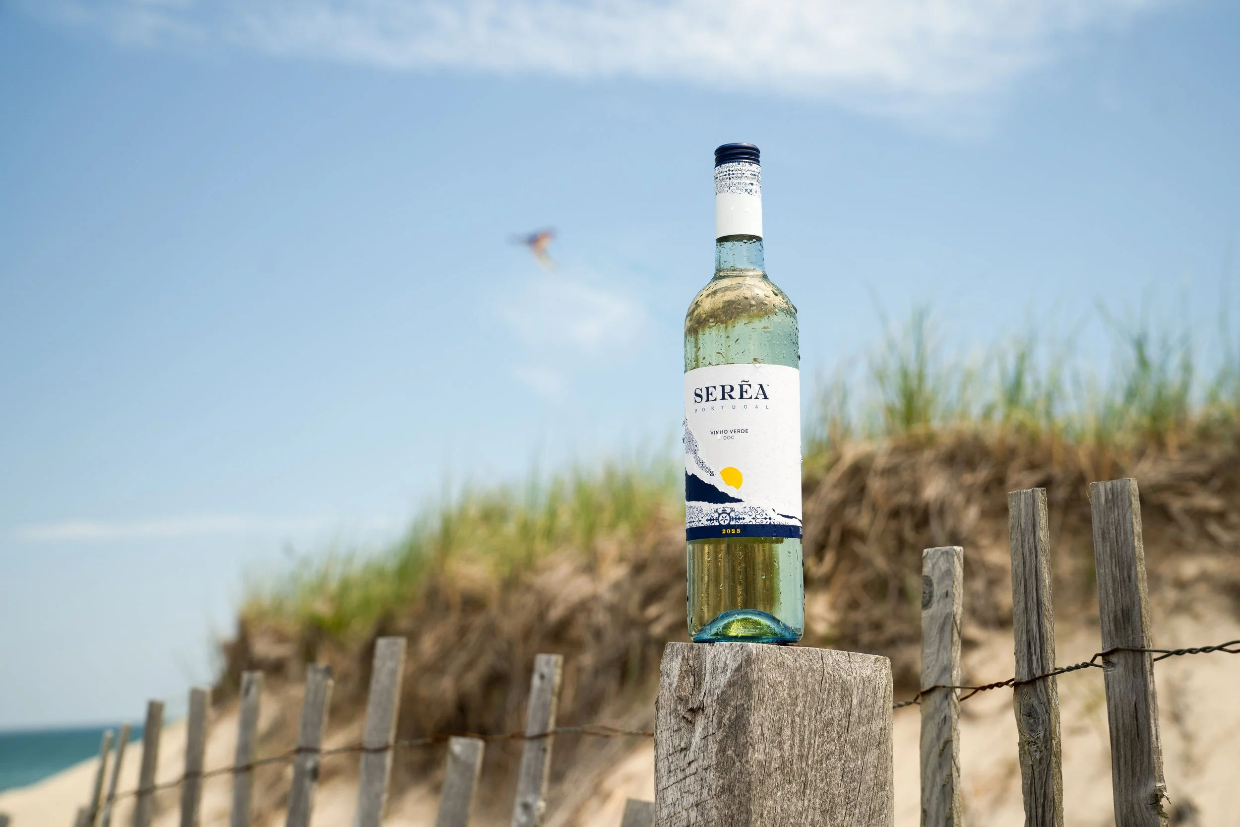 A clear glass bottle labeled Serea Vinho Verde wine with a illustration of a yellow sun and blue ocean waves, placed on a wooden fence post at a beach, with sand dunes and a blue sky in the background.
