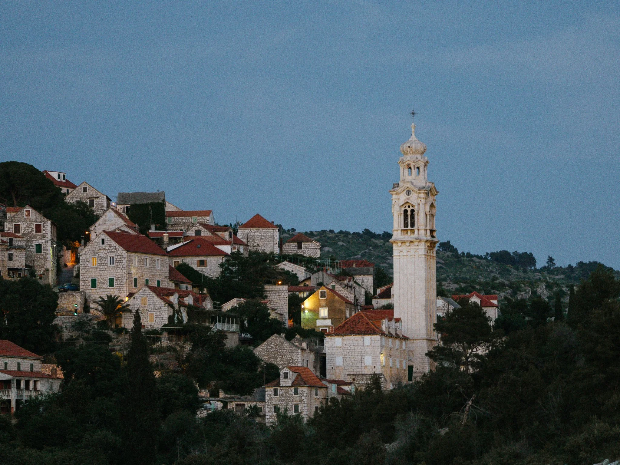 A scenic view of a hillside town in Croatia with white stone houses with red roofs, and a tall white church tower with a clock and a cross on top, under a blue sky.