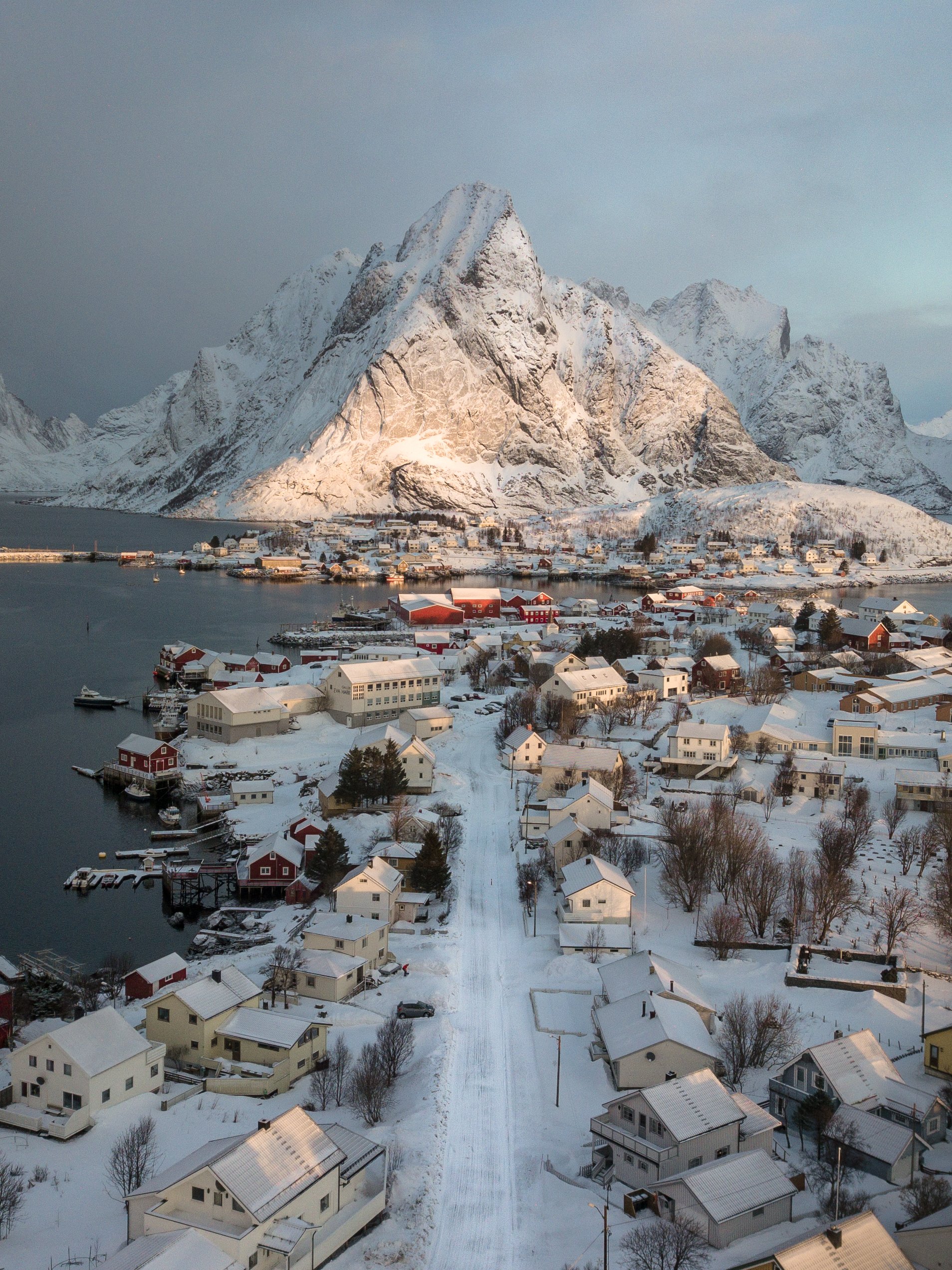A snowy village by a body of water with a large, snow-covered mountain in the background.