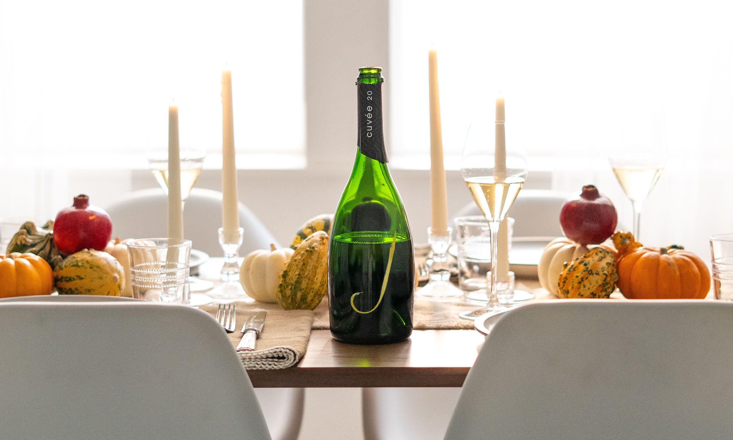 A dining table decorated with pumpkins, gourds, and a bottle of sparkling beverage, set with glasses and candles, in a well-lit room with a large window.