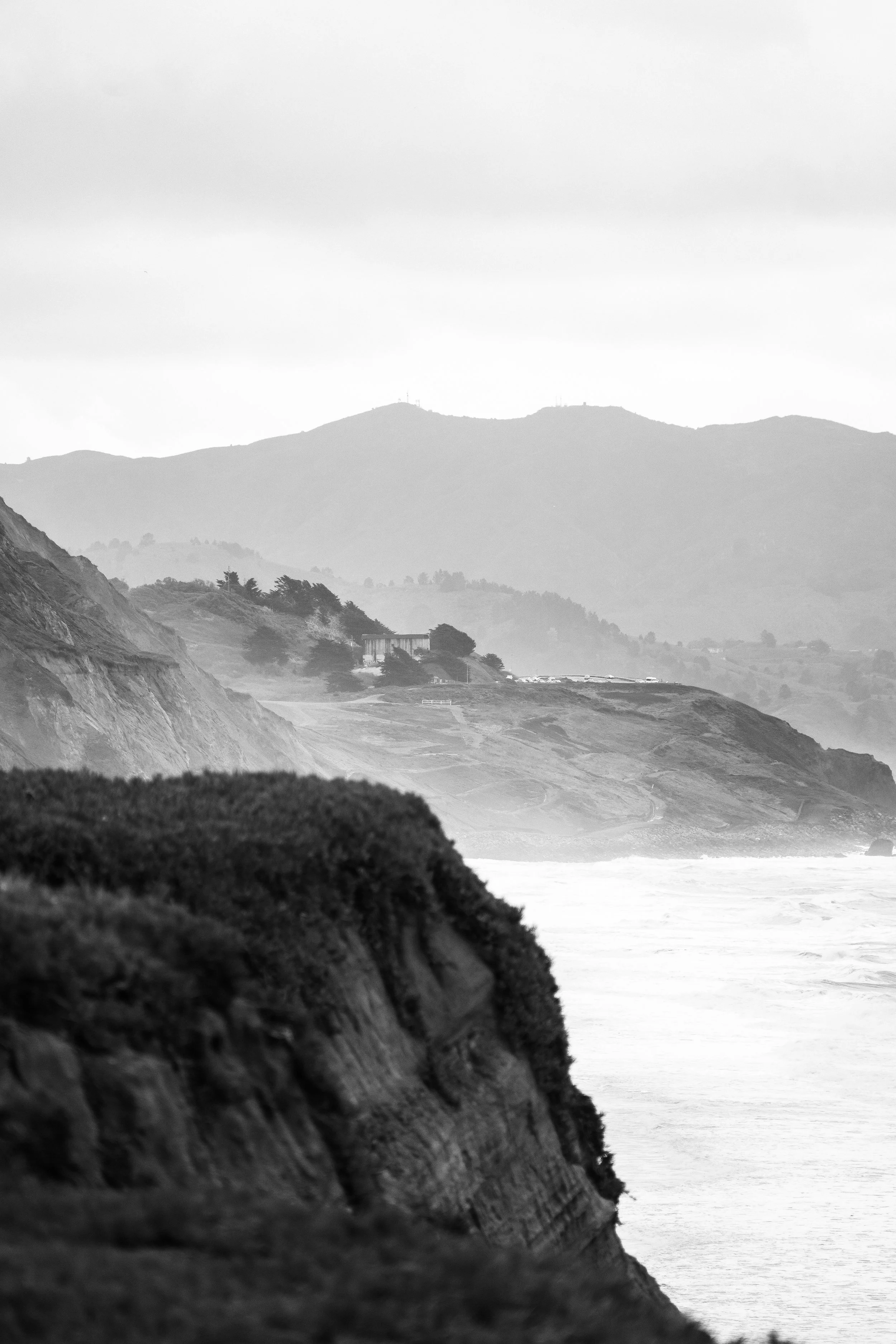 Black and white photo of a coastline with hills and cliffs, water in the foreground, and hills with structures on top in the background.