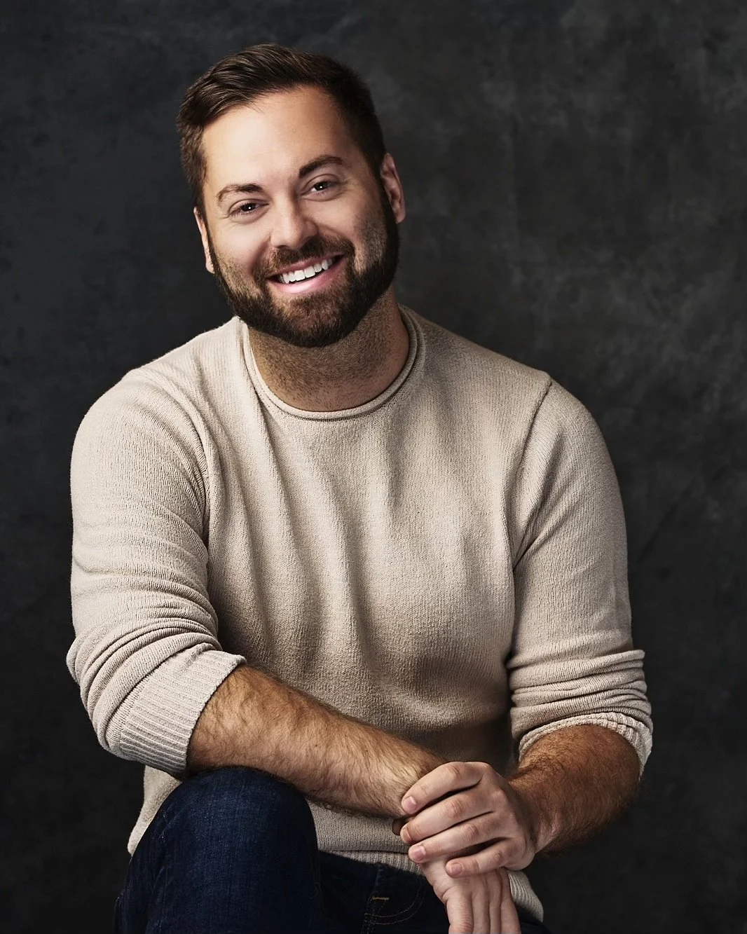 A smiling man with a beard, wearing a beige sweater, seated against a dark textured background.