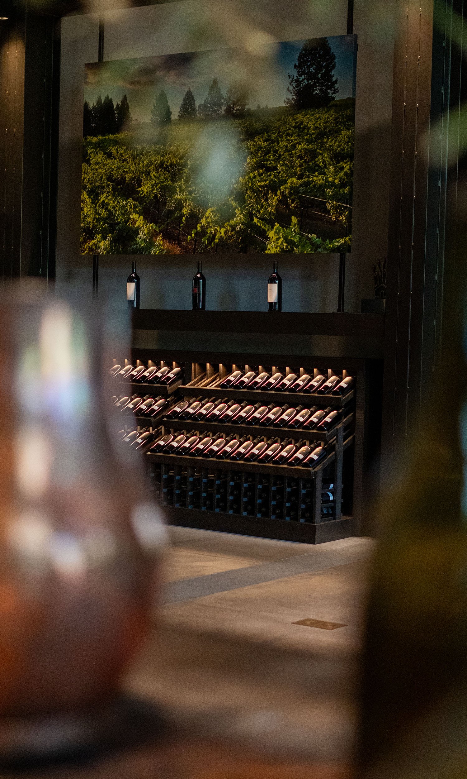 A wine cellar with a rack filled with bottles at the bottom, a countertop with upright wine bottles in the middle, and a landscape painting of a vineyard scene on the wall behind.