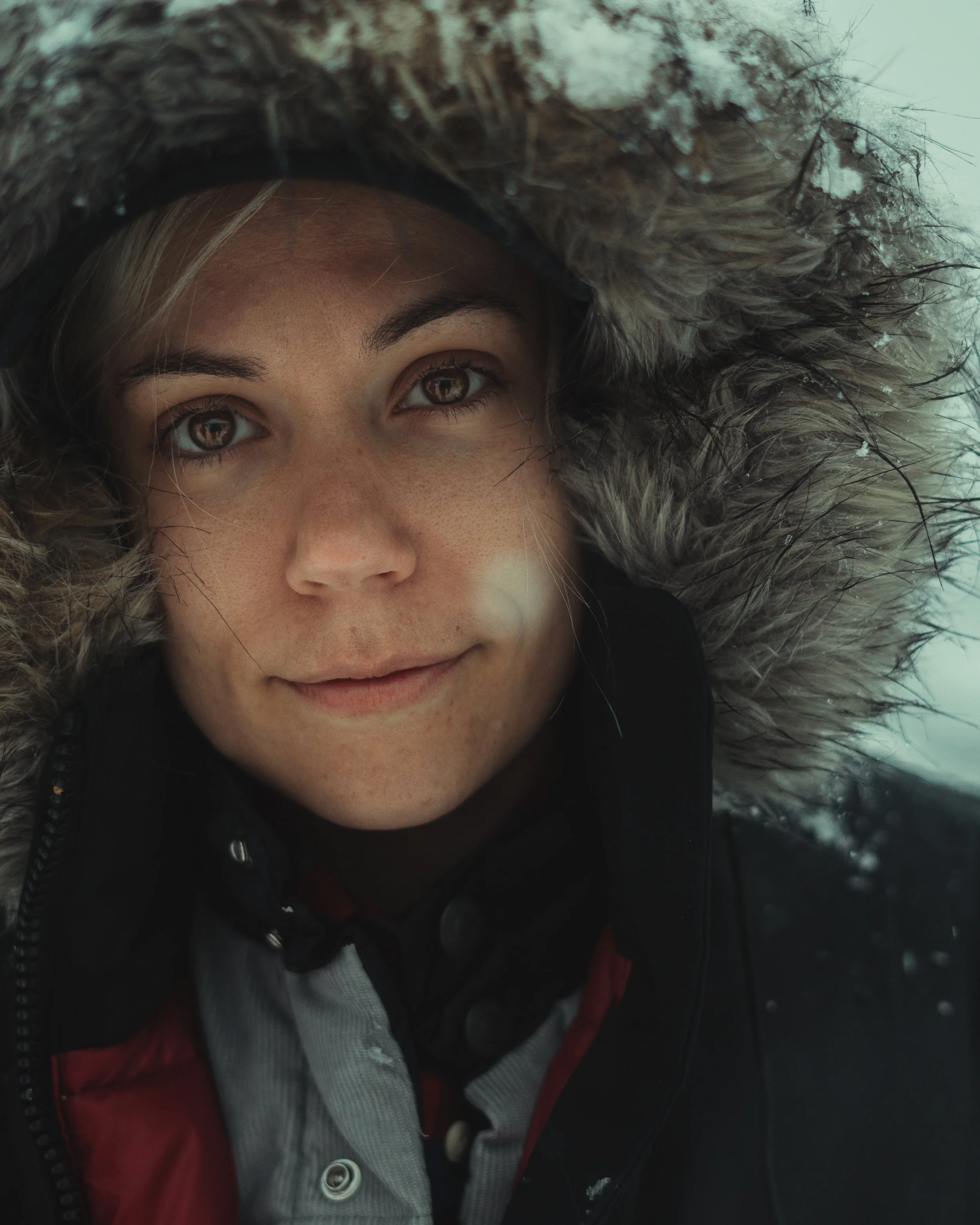 Close-up of a person's face wearing a hood with fur, snow falling.