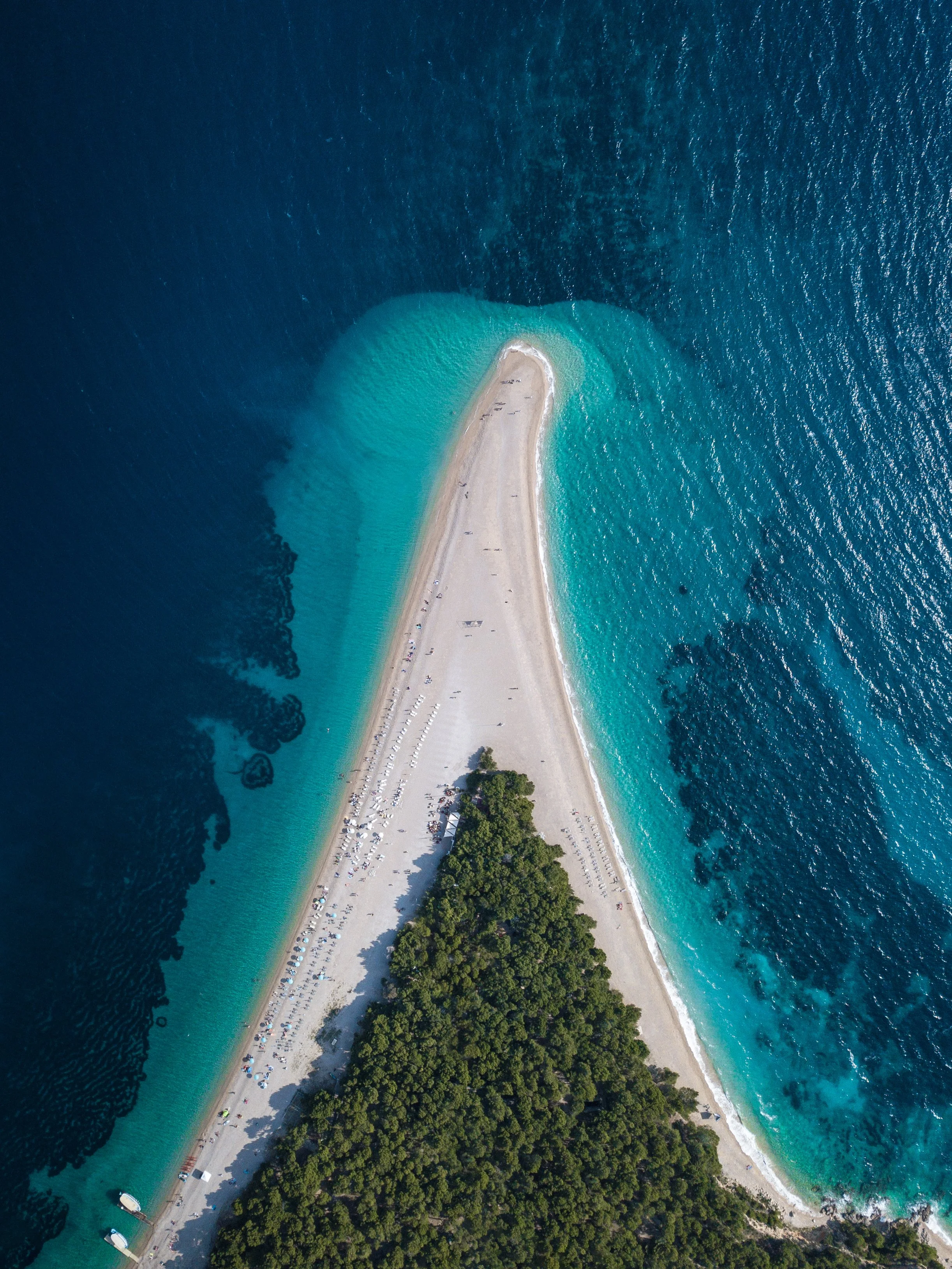 Aerial view of a narrow strip of sandy beach with trees, surrounded by turquoise water, with boats and umbrellas along the shore.