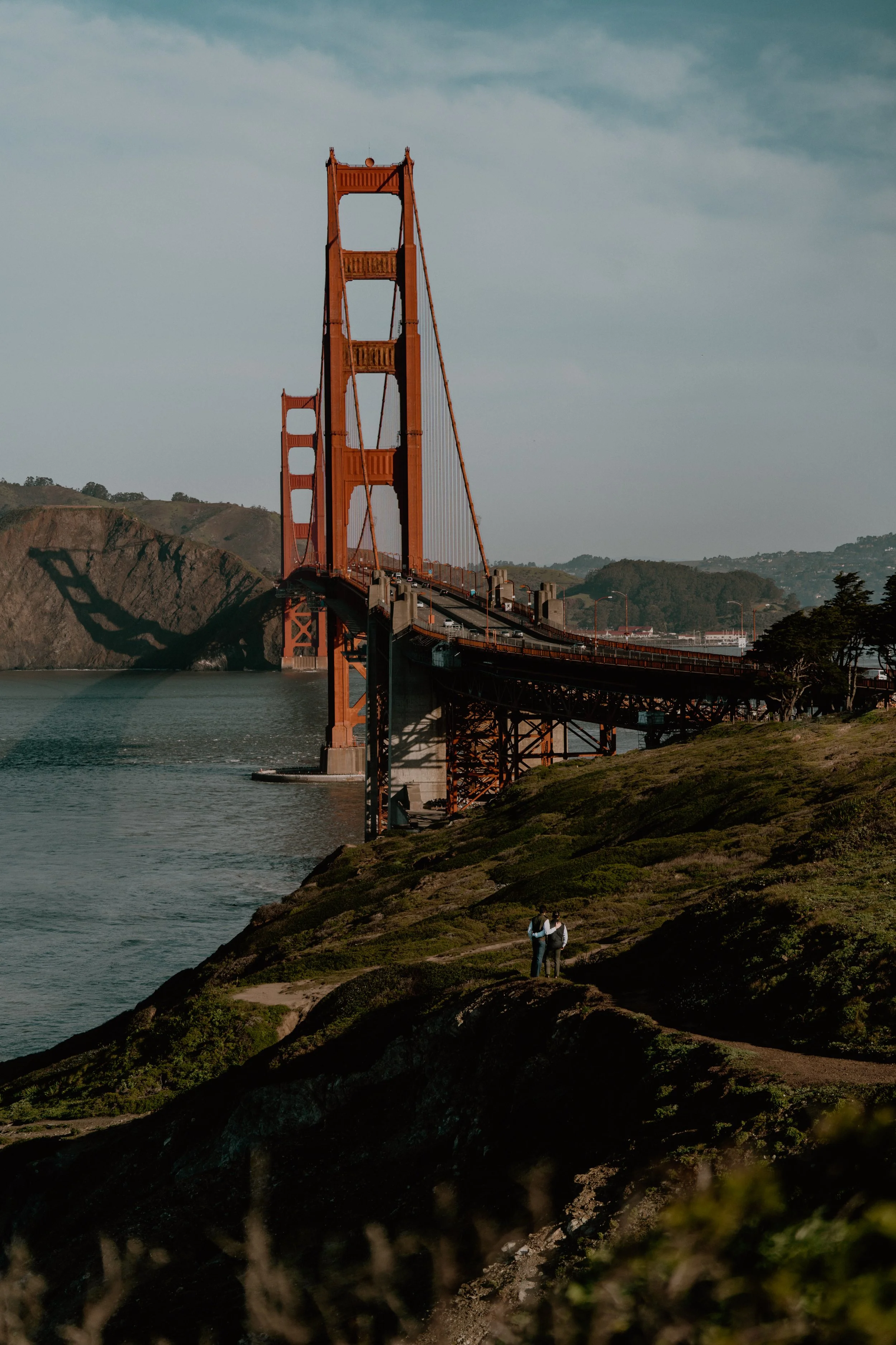 The Golden Gate Bridge in San Francisco during late afternoon or early evening with a couple standing on a hill looking at it.