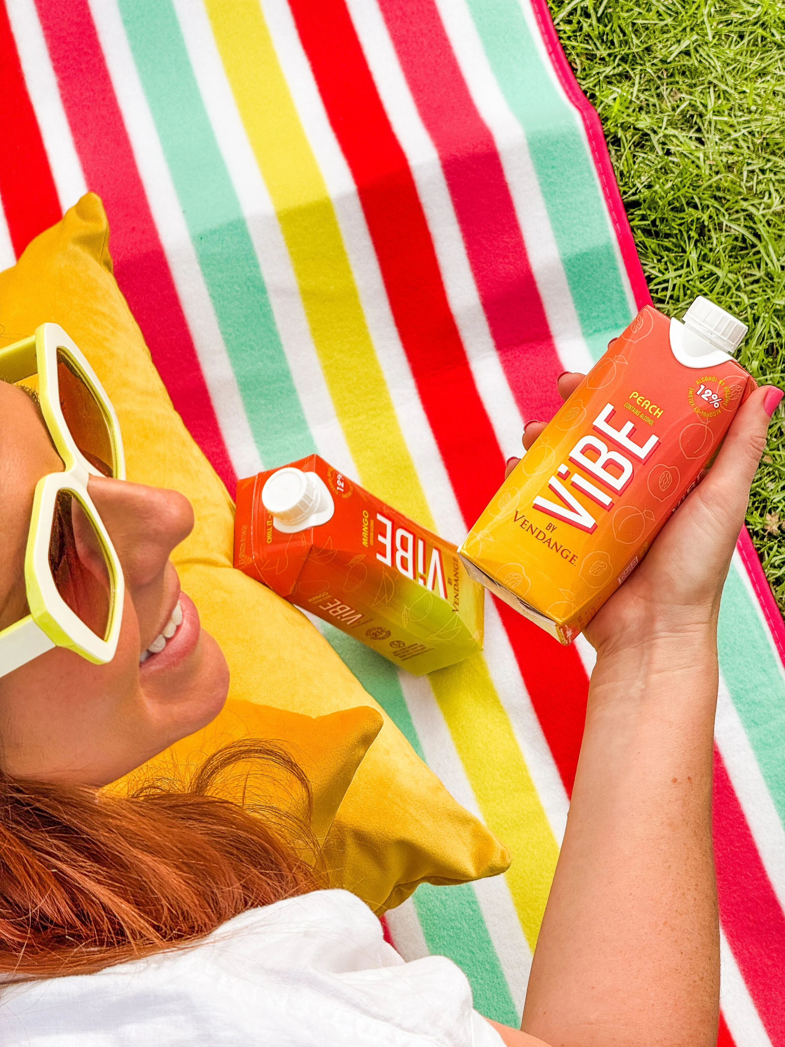 Person with red hair wearing white sunglasses, a yellow shirt or towel, holding a peach-flavored ViBE Twisted Sips, sitting on a colorful striped towel on the grass.