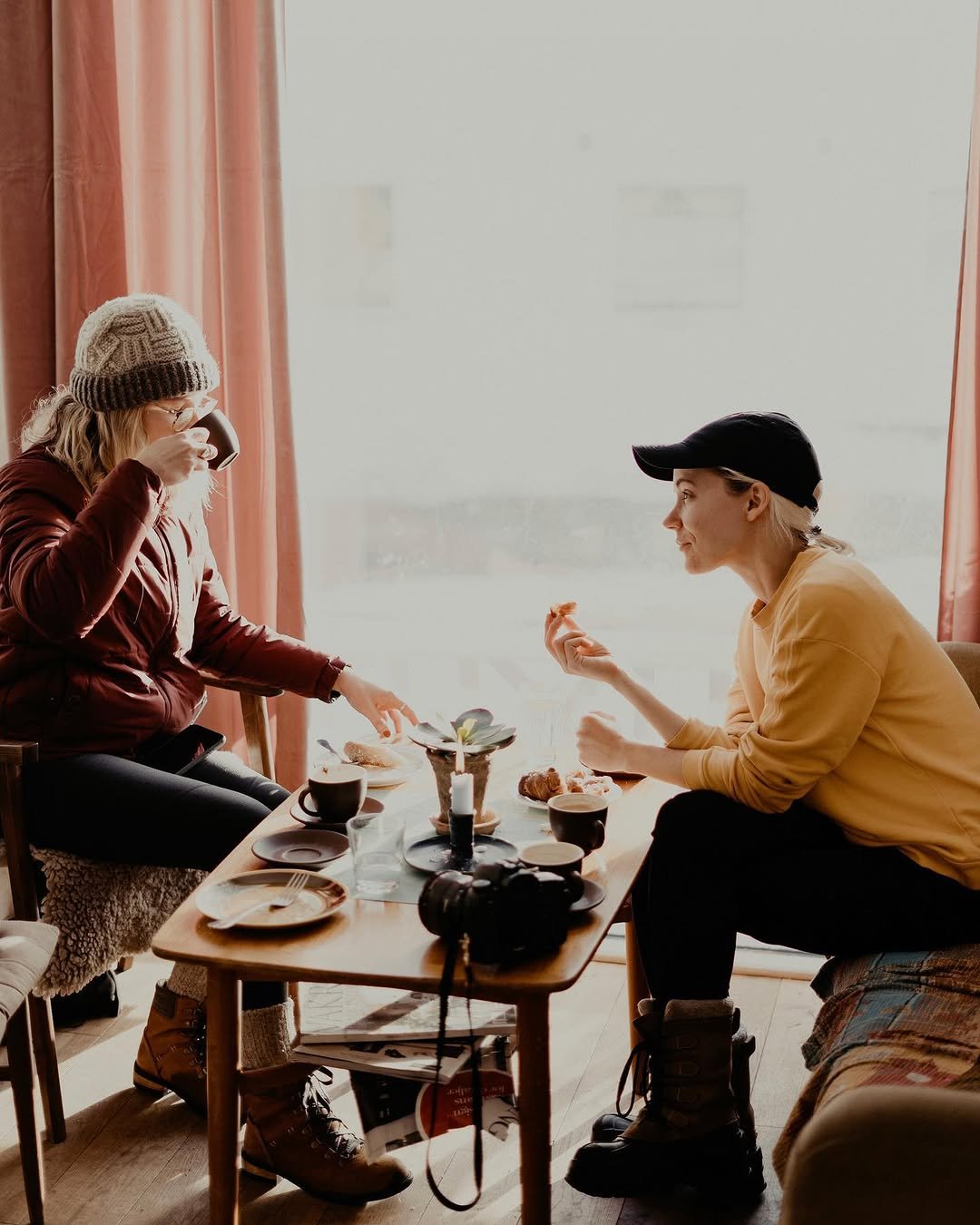 Two women sitting at a table by a large window, sharing tea and snacks. One woman is wearing a knit hat and glasses, drinking from a mug, while the other wears a baseball cap and is holding a snack.