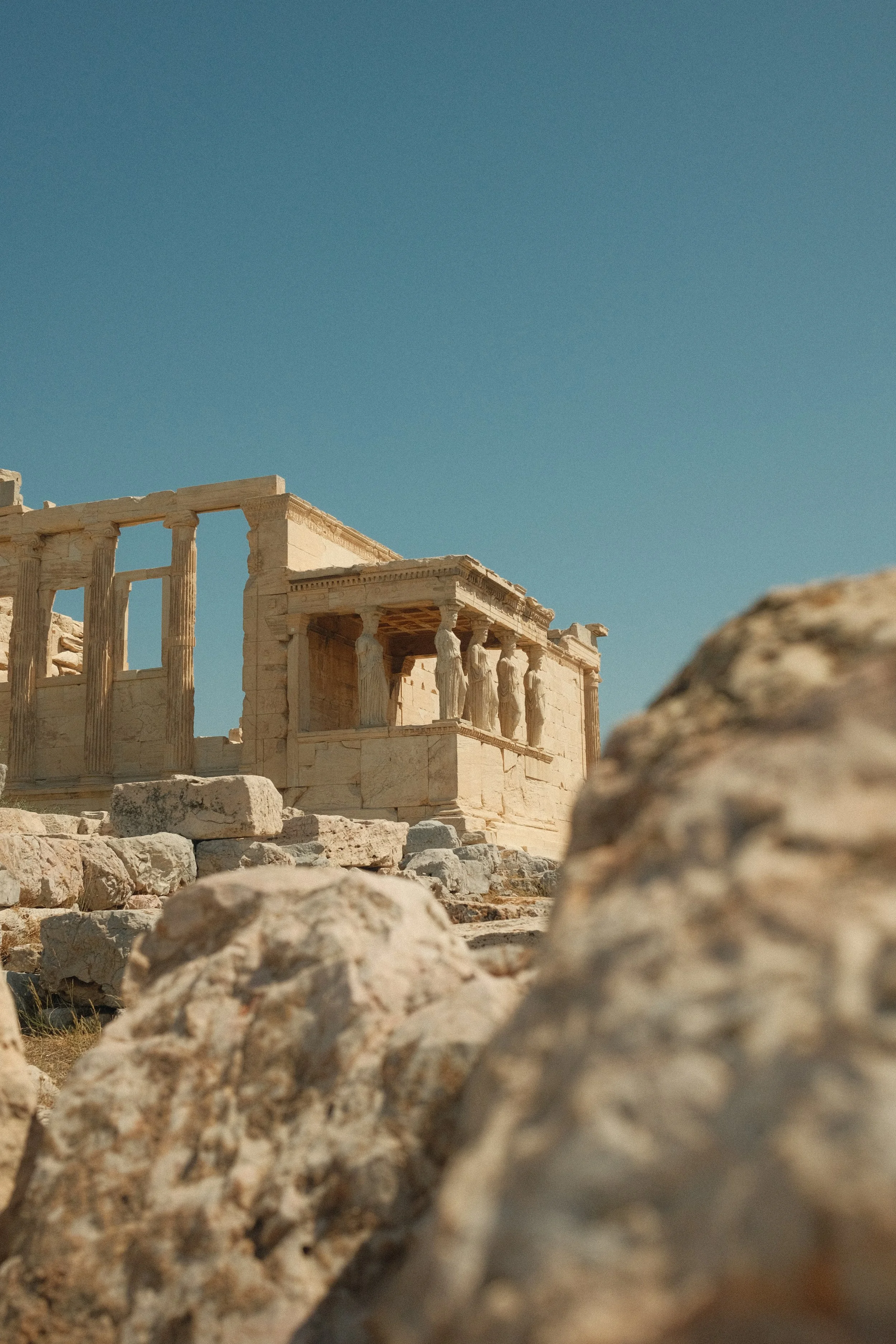 Ancient Greek ruins with columns and structures in ruins, under a clear blue sky, with scattered rocks in the foreground.