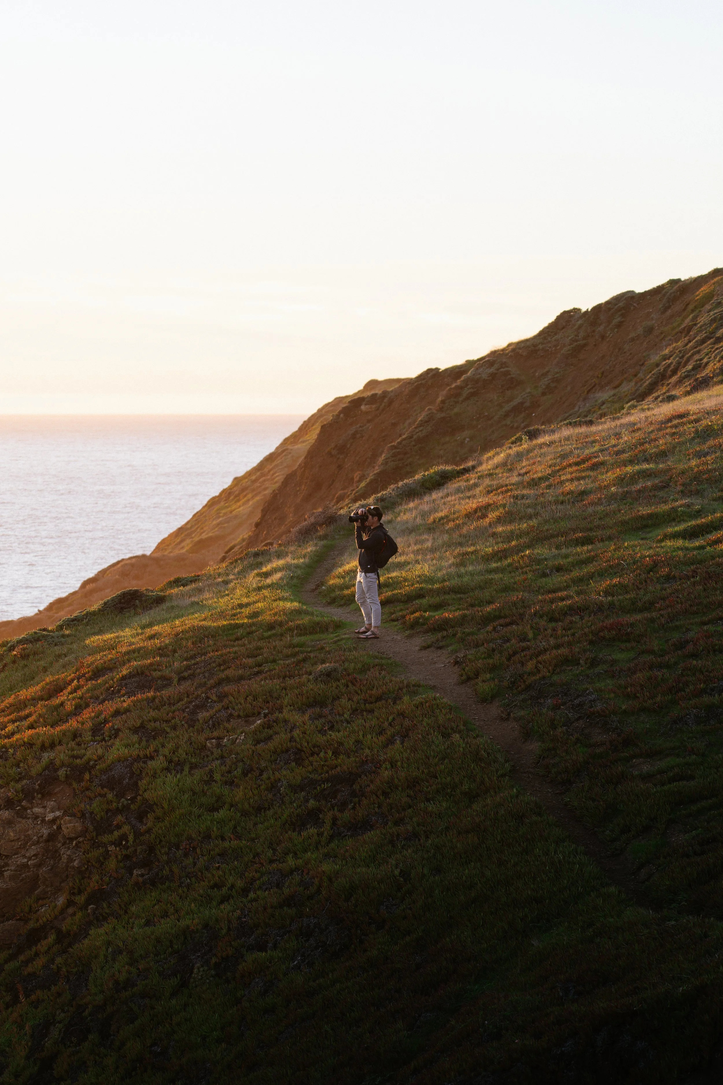 A person with a backpack taking a photograph on a grassy hillside at sunset, overlooking the ocean with cliffs in the distance.