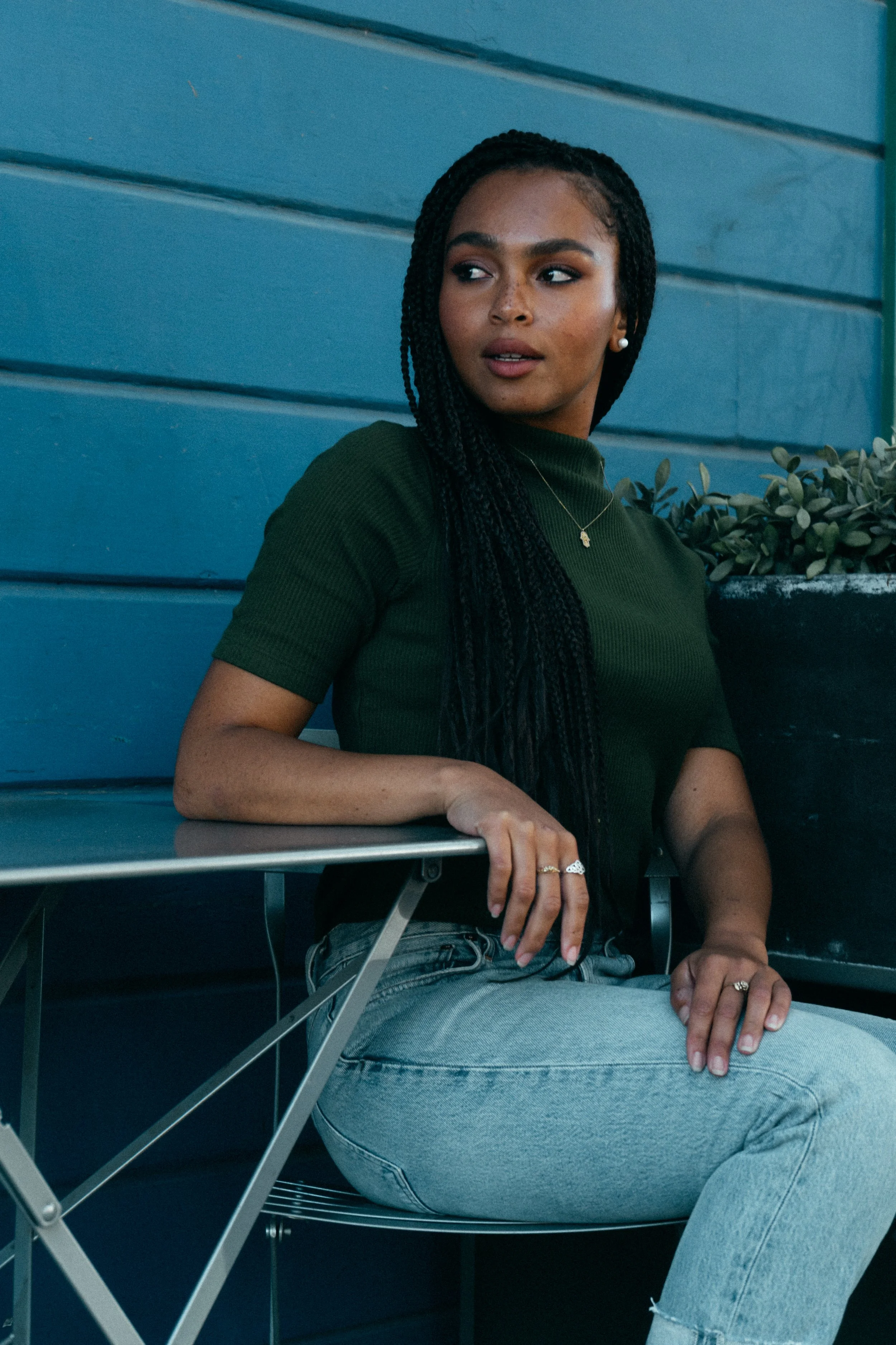 A woman with braided hair, wearing a dark green top and grey jeans, sitting against a blue wall, holding a metal chair with one hand, and looking to her left.