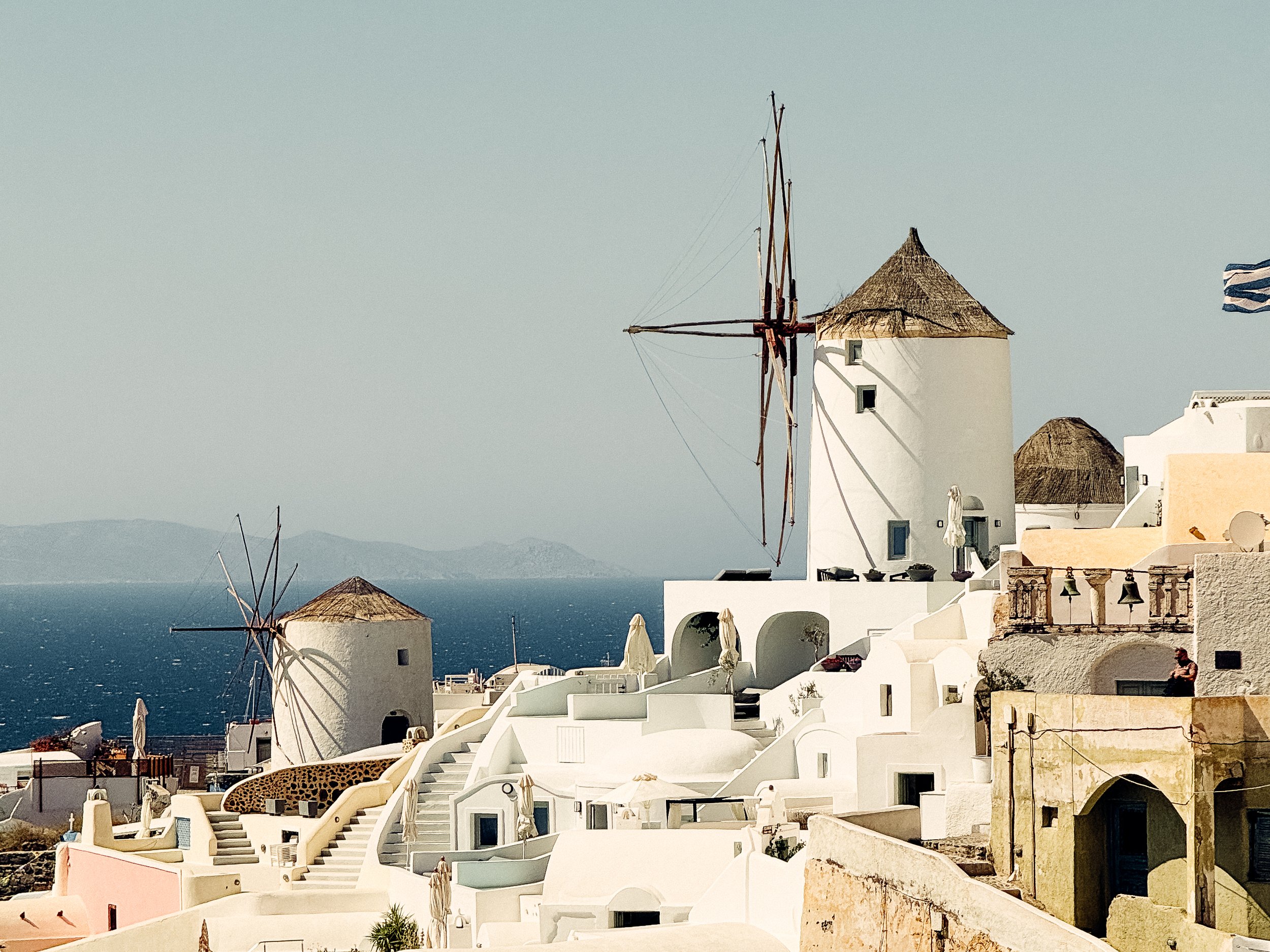 White buildings with rounded edges and terracotta roofs, windmill with wooden blades, Greek flag, and a view of the sea in the background.