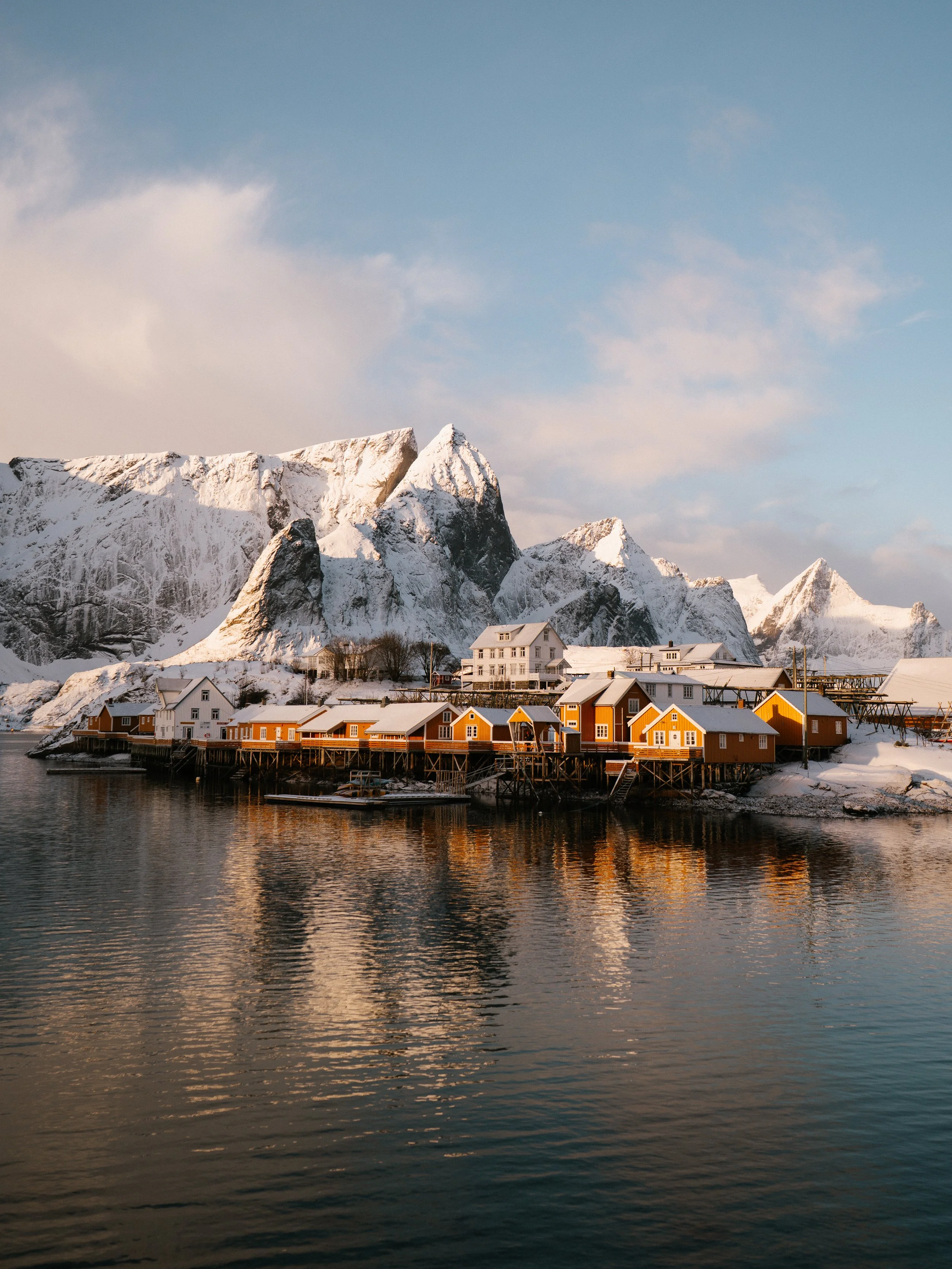 Colorful houses on stilts along a waterfront with snow-covered mountains in the background under a partly cloudy sky.